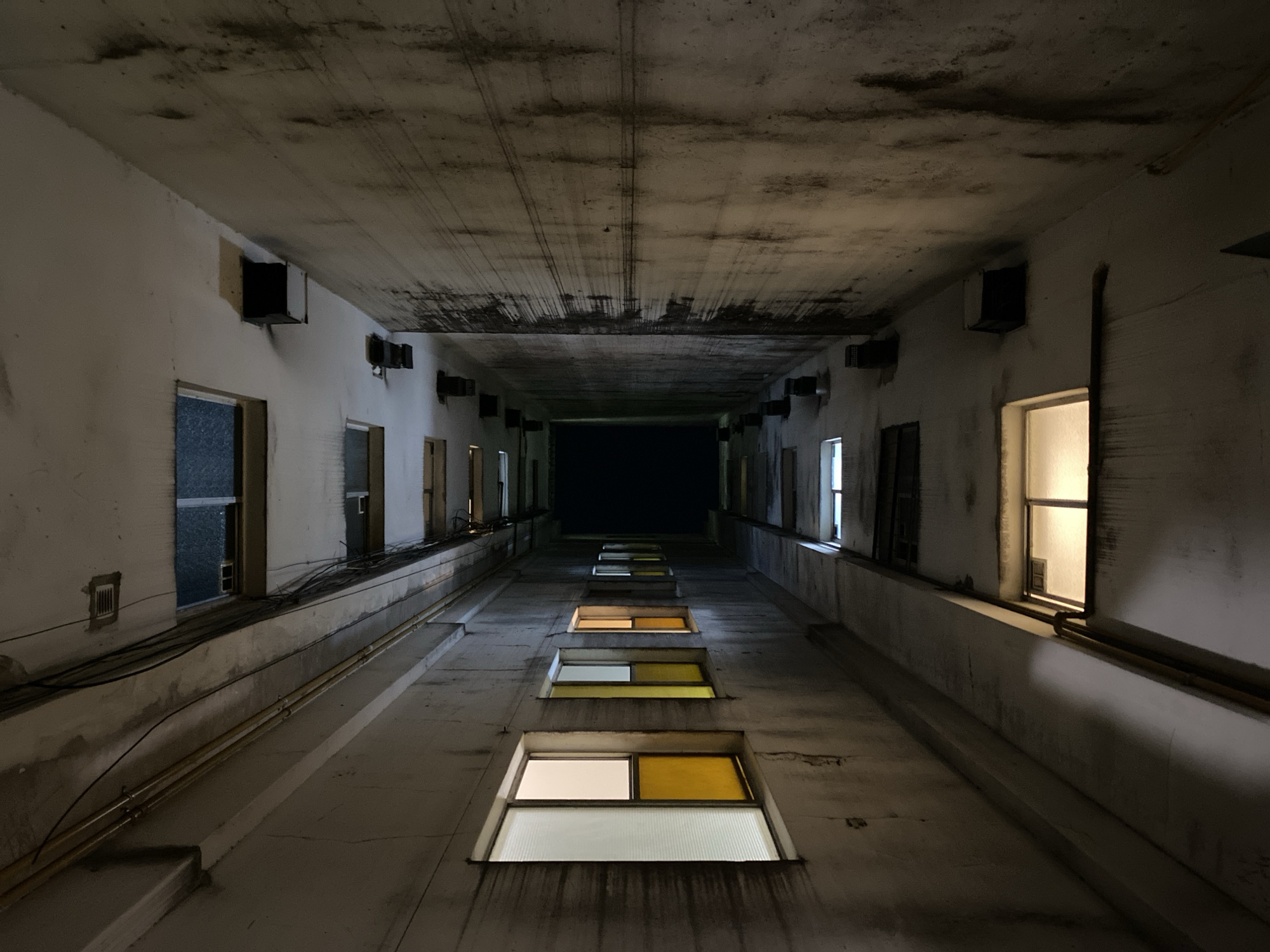 The image depicts a narrow, concrete-walled corridor or shaft, viewed from a low angle looking upwards. The location is not visible, and the location in the city of Buenos Aires is not discernible from the image.
The corridor appears institutional or utilitarian, with minimal ornamentation. The walls are stained and marked, suggesting age or neglect. A series of windows are arrayed along one side of the corridor, some illuminated from within with a warm, yellow light. The far end of the corridor is dark, possibly indicating an opening or a lack of light.
Electrical conduits or pipes run along the walls. There are no people visible. The perspective creates a sense of depth and confinement.