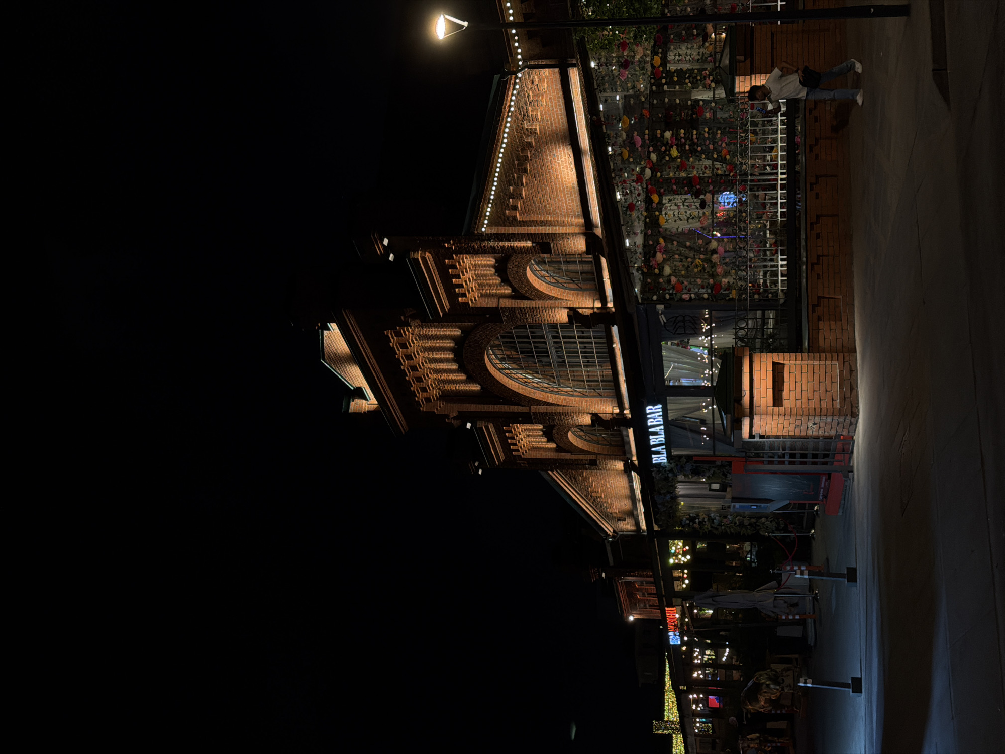 The image shows the exterior of the Bla Bla Bar, a restaurant located in Moscow, Russia. The bar is a brick building with large windows and a neon sign. It is decorated with flowers. The sky is black and there are people walking on the street.