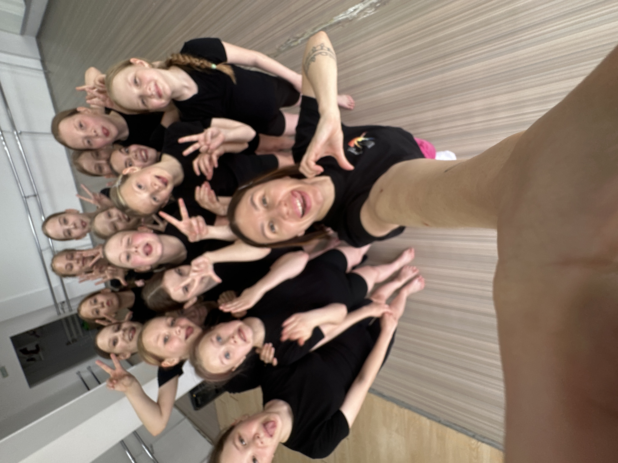 A selfie captured a group of approximately 15 individuals, primarily children, dressed in black outfits. The woman holding the camera is also wearing a black t-shirt and stands at the center of the group. Many of the children are flashing peace signs, and some are sticking out their tongues for the photo. The background appears to be a dance studio with light-colored, wood-like flooring. A mirrored wall with a railing runs along the left side of the frame, reflecting the group and adding to the image's depth. The photo seems to be taken indoors, possibly in Votkinsk, Russia, in an exercise or dance setting. The environment appears well-lit, and the overall atmosphere is cheerful and active.