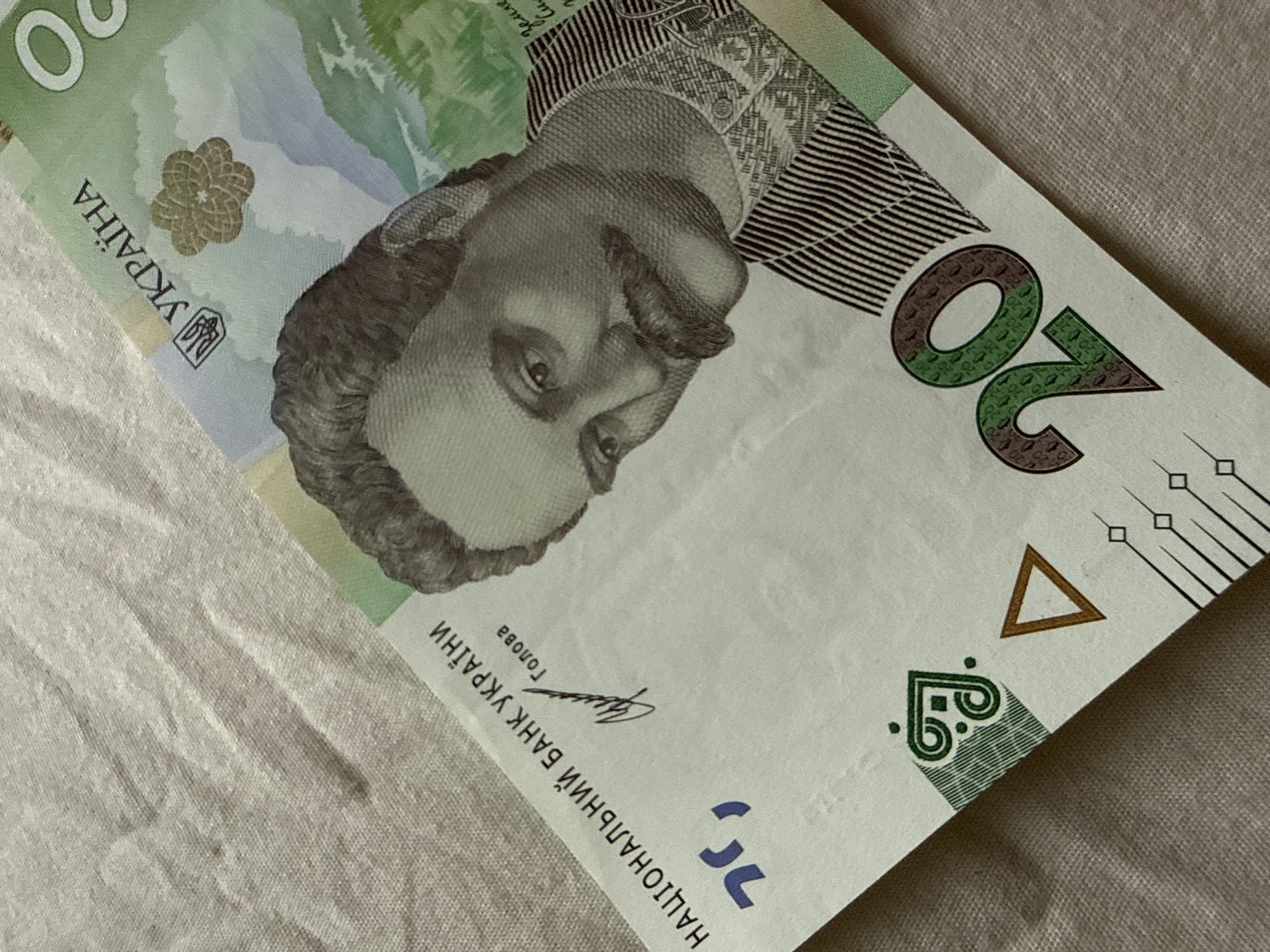 The image shows a close-up of a Ukrainian 20 hryvnia banknote. The banknote is partially laid out on a textured, light-colored fabric surface, which appears to be a tablecloth or a similar textile. The banknote is angled diagonally across the frame.
Prominent features visible on the banknote include:
- A portrait of Ivan Franko, a Ukrainian writer and political figure, is visible in the center. He has a mustache and is depicted in a suit.
- The text "НАЦІОНАЛЬНИЙ БАНК УКРАЇНИ" (National Bank of Ukraine) is printed on the left side.
- The number "20" is prominently displayed in the lower right corner.
- Ukrainian national symbols and floral motifs are also present.
- The background of the banknote depicts a landscape.
The fabric surface on which the banknote rests has a fine, woven texture and is creased in some areas, suggesting it is soft and pliable. The lighting in the image is soft, casting gentle shadows.
There is no indication of the city of Bad Sachsa, Germany, in the image. The image solely focuses on the banknote and the surface it is on.