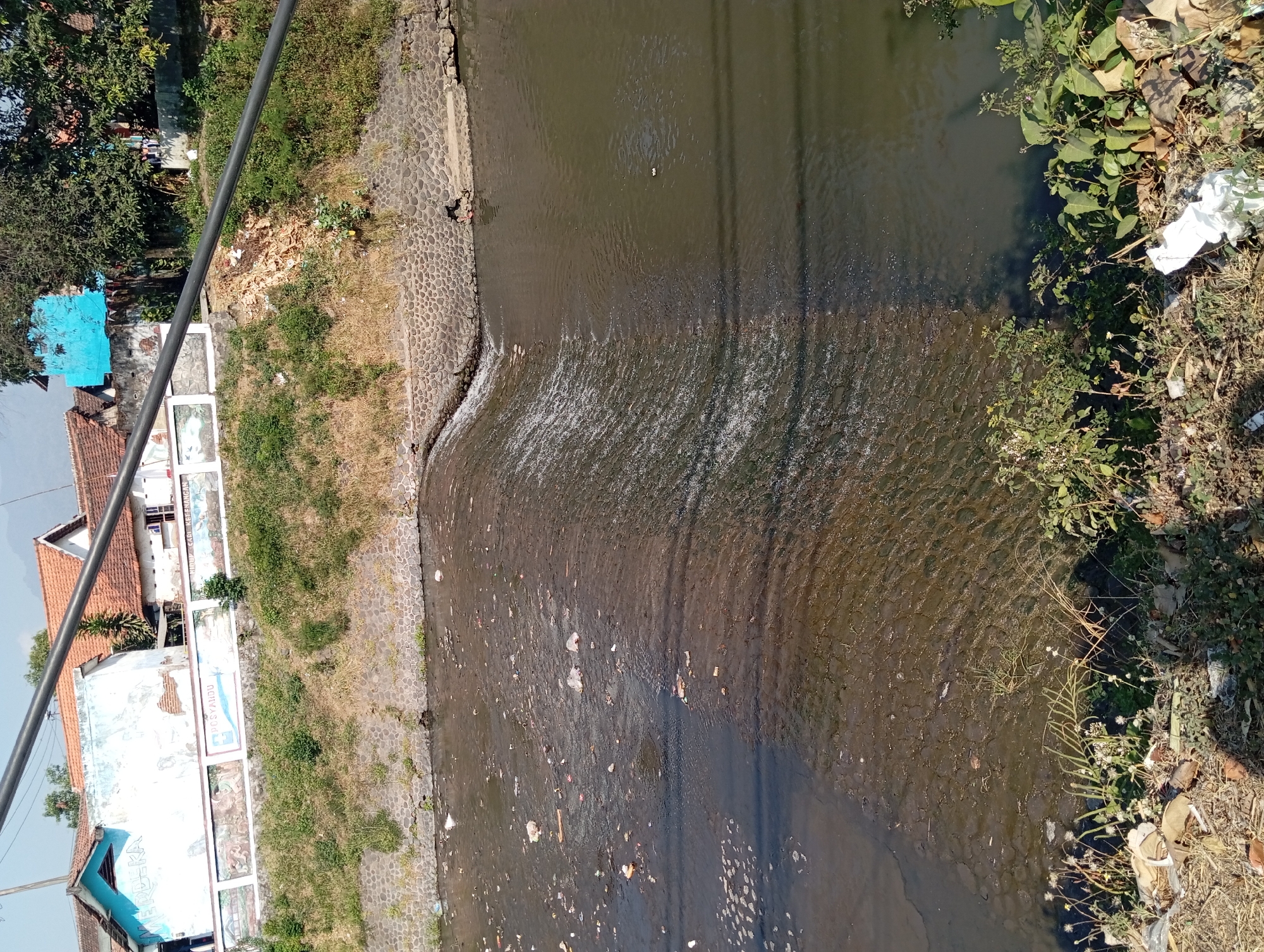 The image shows a polluted river flowing through Kepulungan, Indonesia. The river is filled with trash, including plastic bottles, bags, and other debris. The banks of the river are lined with vegetation and some houses are visible in the background. A small waterfall is present in the river, with water flowing over a series of rocks. The scene depicts environmental degradation and the impact of human activity on natural resources.