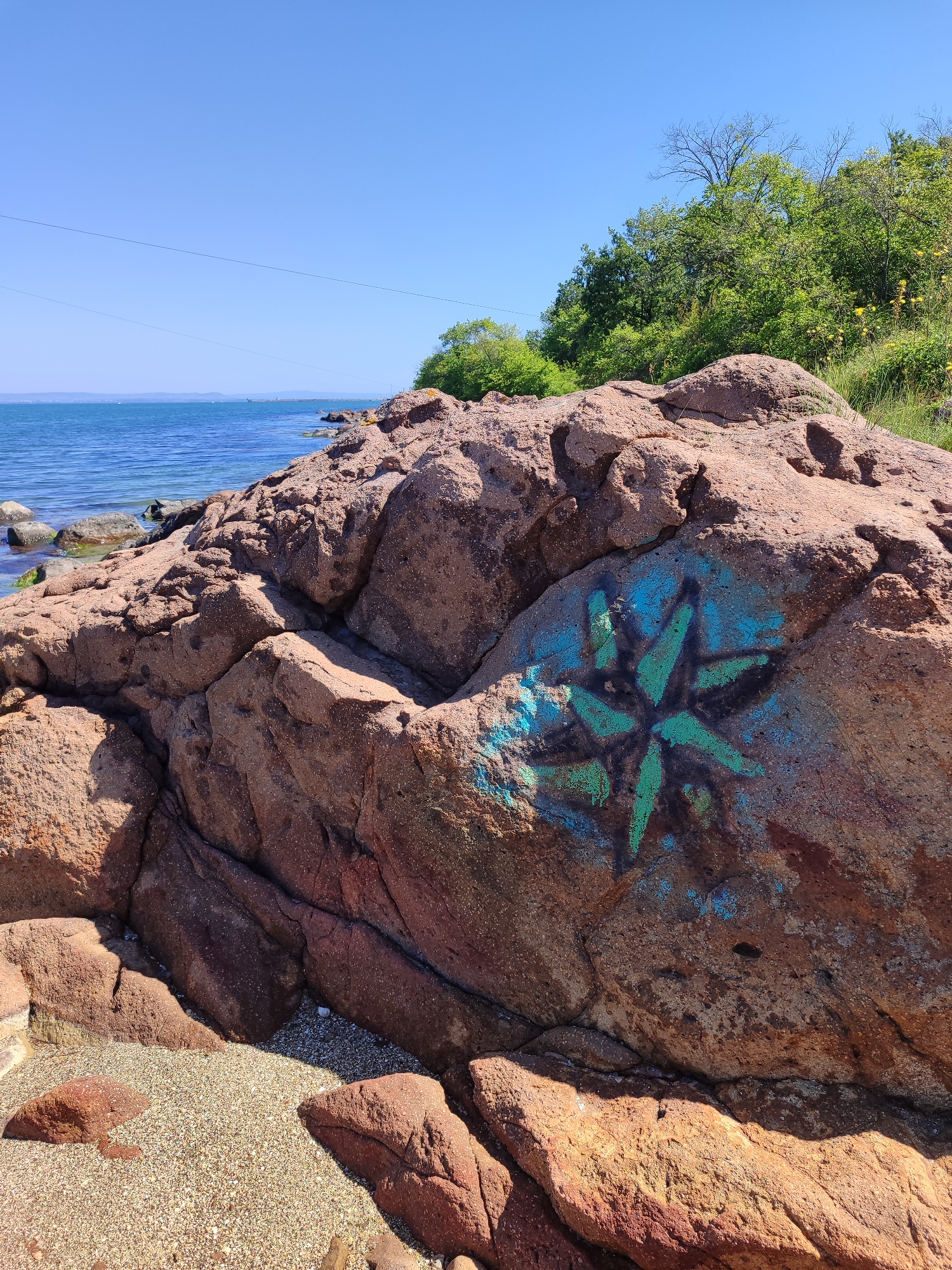A sunny, outdoor scene depicts a large, reddish-brown rock formation on a pebble beach along the coastline. The sea and a strip of land are visible in the background beneath a clear blue sky. Part of a tree line extends on the coastline. Utility wires are visible stretching overhead. A star-shaped design is painted in teal and black paint on one face of the rock formation. Small, white and tan pebbles make up the beach in the foreground. The photograph was taken in Burgas, Bulgaria.