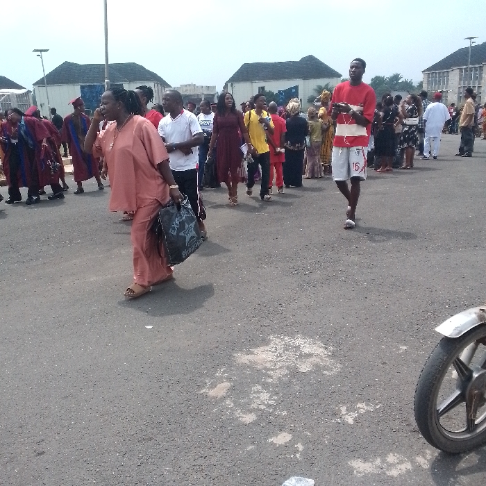 The scene depicts an outdoor gathering of numerous individuals on a paved area in OGBOKO, Nigeria. The ground is asphalt with scattered light-colored patches. The sky is overcast. In the background, several multi-story buildings with dark, sloped roofs are visible, interspersed with sparse vegetation and tall utility poles.
In the foreground, slightly left of center, a dark-skinned adult female walks towards the right. She wears a peach-colored loose-fitting top and matching trousers with light brown open-toed sandals. Her left hand is raised near her face, and her right hand holds a dark reusable shopping bag with white lettering. To her left, another dark-skinned adult male in a white t-shirt and dark trousers walks in the same direction. Further right, in the mid-ground, a dark-skinned adult male walks towards the left, wearing a red short-sleeved t-shirt with white stripes, white athletic shorts with the number "16", and white flip-flops. He is using a smartphone held at chest level and has white earbuds in his ears.
Numerous other individuals are present throughout the scene, walking in various directions. Many are dressed in diverse attire, including some in dark academic gowns with colored stoles or hoods, others in traditional Nigerian clothing, and some in casual wear. A metallic front wheel and fender of a motorcycle or scooter are visible in the bottom right corner of the frame.