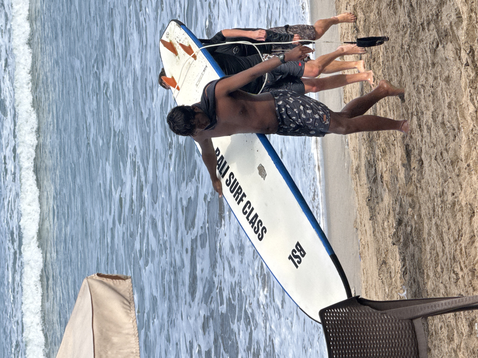 In Kuta, Indonesia, on a sandy beach with the ocean in the background, three individuals are walking with a surfboard. The primary subject, a man in the foreground, carries the surfboard on his left shoulder. He is wearing dark patterned swim shorts and has a dark cloth draped around his neck. To his right, two other individuals, partially obscured, also appear to be carrying or assisting with the surfboard. The surfboard is white with blue trim and orange fins. In the far left of the image, a portion of a beige umbrella is visible. The ocean features gentle waves breaking on the shore, and the sand is a light brown.