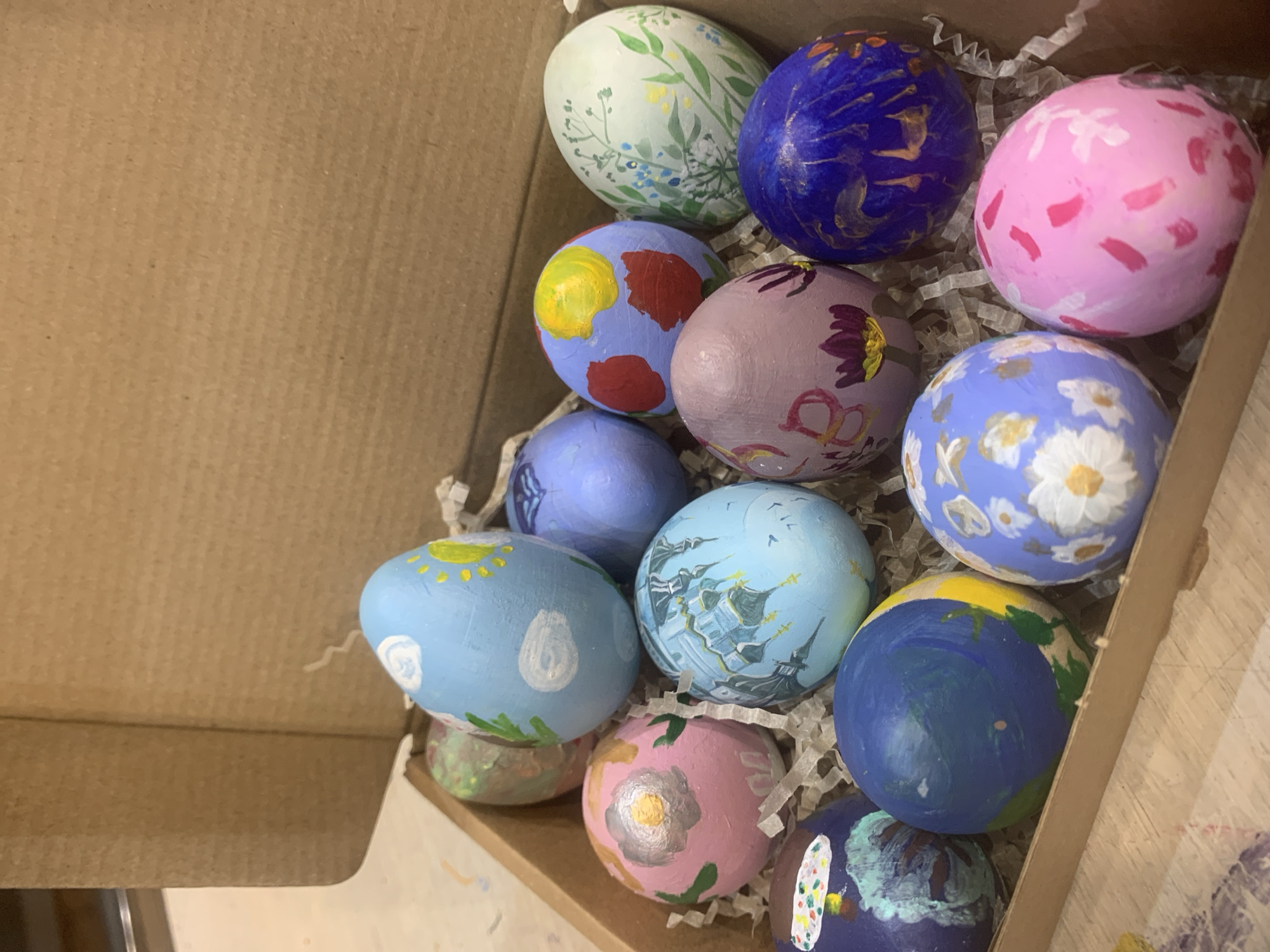 A cardboard box contains thirteen hand-painted Easter eggs nestled in white shredded paper. The eggs display a variety of designs, including flowers, landscapes, celestial bodies, and abstract patterns. The scene is lit with natural light and appears to be indoors. There is no indication of Izhevsk, Russia.