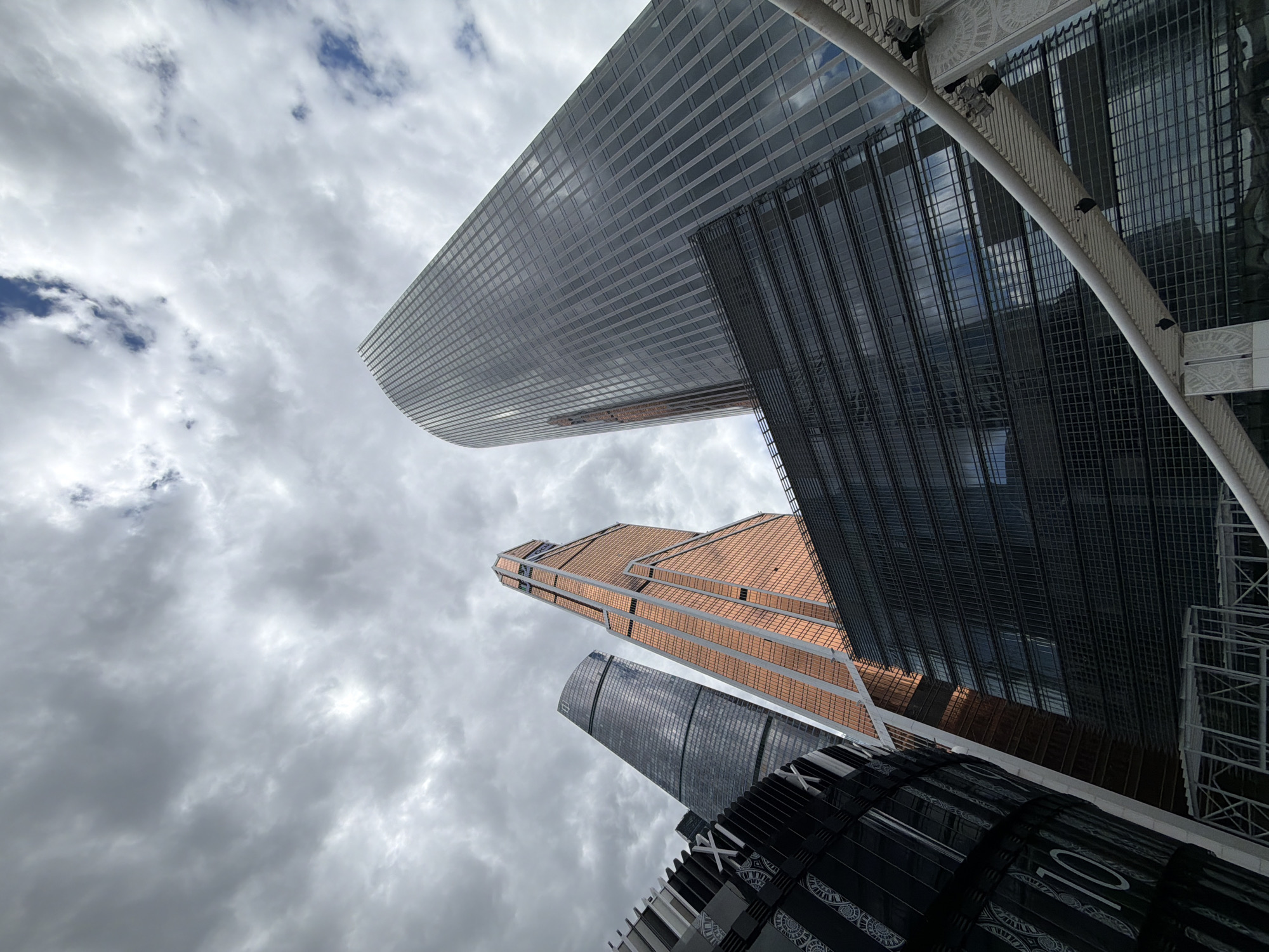 The image captures a low-angle view of several modern skyscrapers in Moscow, Russia. The architecture is characterized by sleek, geometric designs with glass and metal facades. A cloudy sky forms the backdrop, with shades of gray and hints of blue. The buildings exhibit various textures and reflections due to their glass surfaces. The composition emphasizes the height and scale of the structures, creating a sense of urban grandeur.