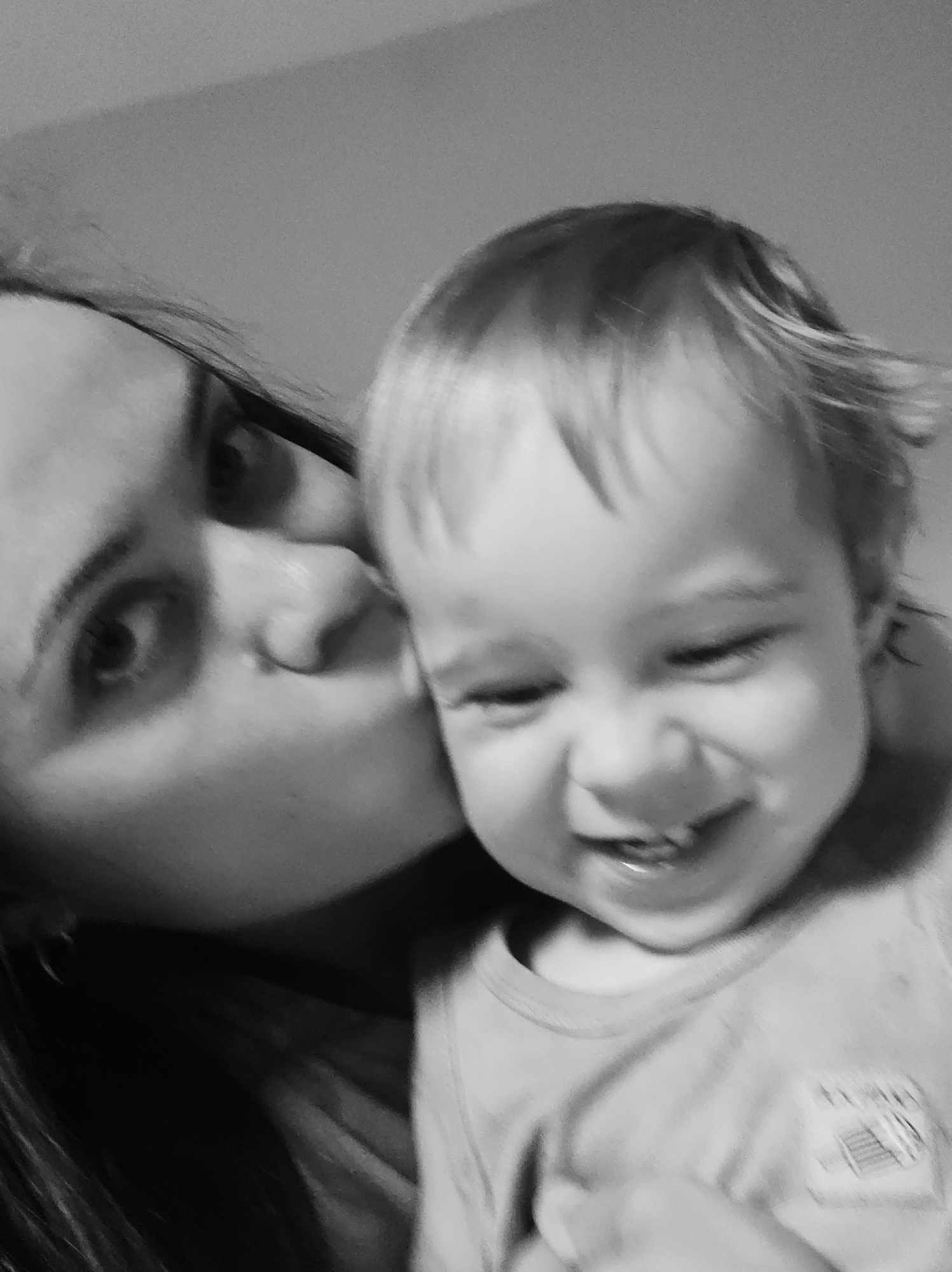 A monochrome, soft-focus image depicts an adult and a young child in close proximity. The adult, positioned on the left, has visible dark hair and a left eye directed towards the camera. The right side of the adult's face, including the cheek and mouth, is pressed against the upper right side of the child's head, suggesting a close physical contact. The child, a toddler with light-colored hair, occupies the right frame. The child's eyes are mostly closed due to a wide smile, revealing visible teeth. The child wears a light-colored top with an indistinct pattern or emblem on the right chest area. The background is a plain, uniformly grey, out-of-focus surface, indicating an indoor setting with soft, ambient lighting. The scene occurs in an unspecified indoor location in Unknown, Unknown.