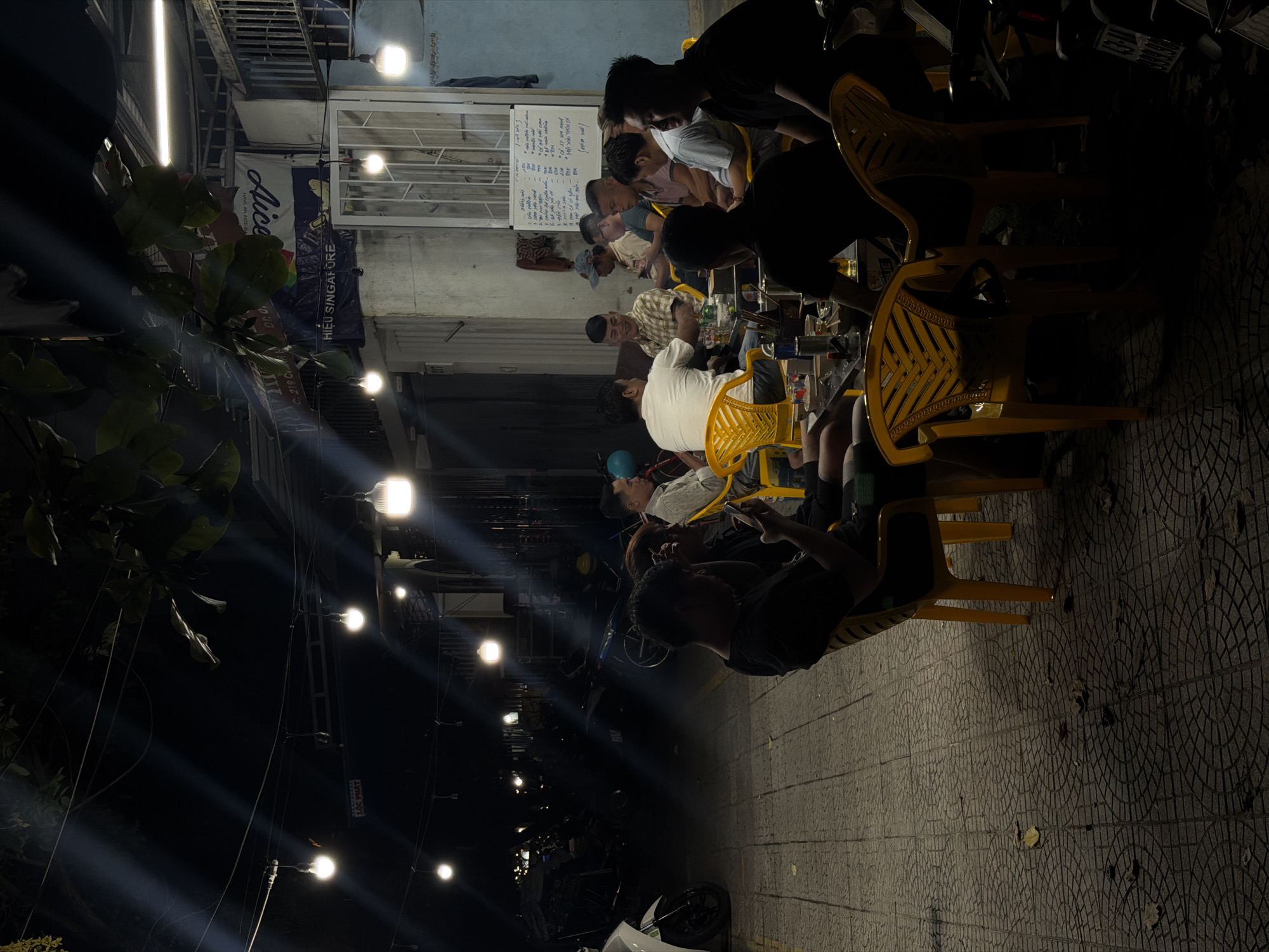 At night in Đà Nẵng, Vietnam, people sit at plastic tables and chairs alongside a street lit by overhead lights. The street is paved with interlocking circular patterned tiles. There are parked scooters. Overhead signage reads "Alice" and "Hiệu Singapore." A menu is affixed to a wall. People are seated at tables. Some are looking at phones.