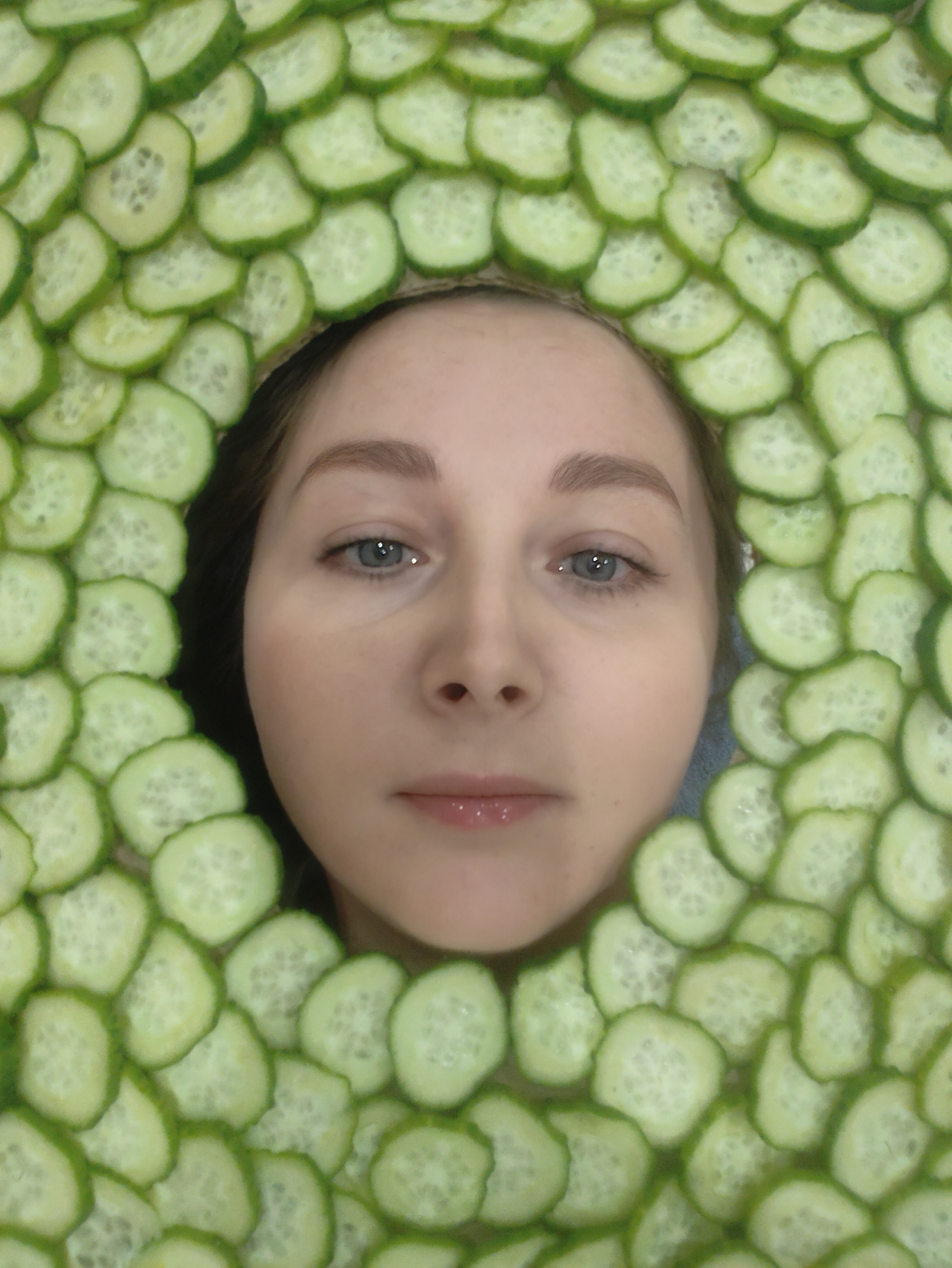 A fair-skinned woman with light brown hair is centered in the frame, surrounded by overlapping slices of cucumbers. Her face, featuring light blue eyes, a straight nose, and full lips with a slight sheen, is the focal point. The cucumbers form a circular arrangement around her face, with slight variations in green hues. The setting is indoors, with the woman possibly lying down or positioned in a specific way to achieve the composition. There is no visible indication of the specific location as the city is unknown.