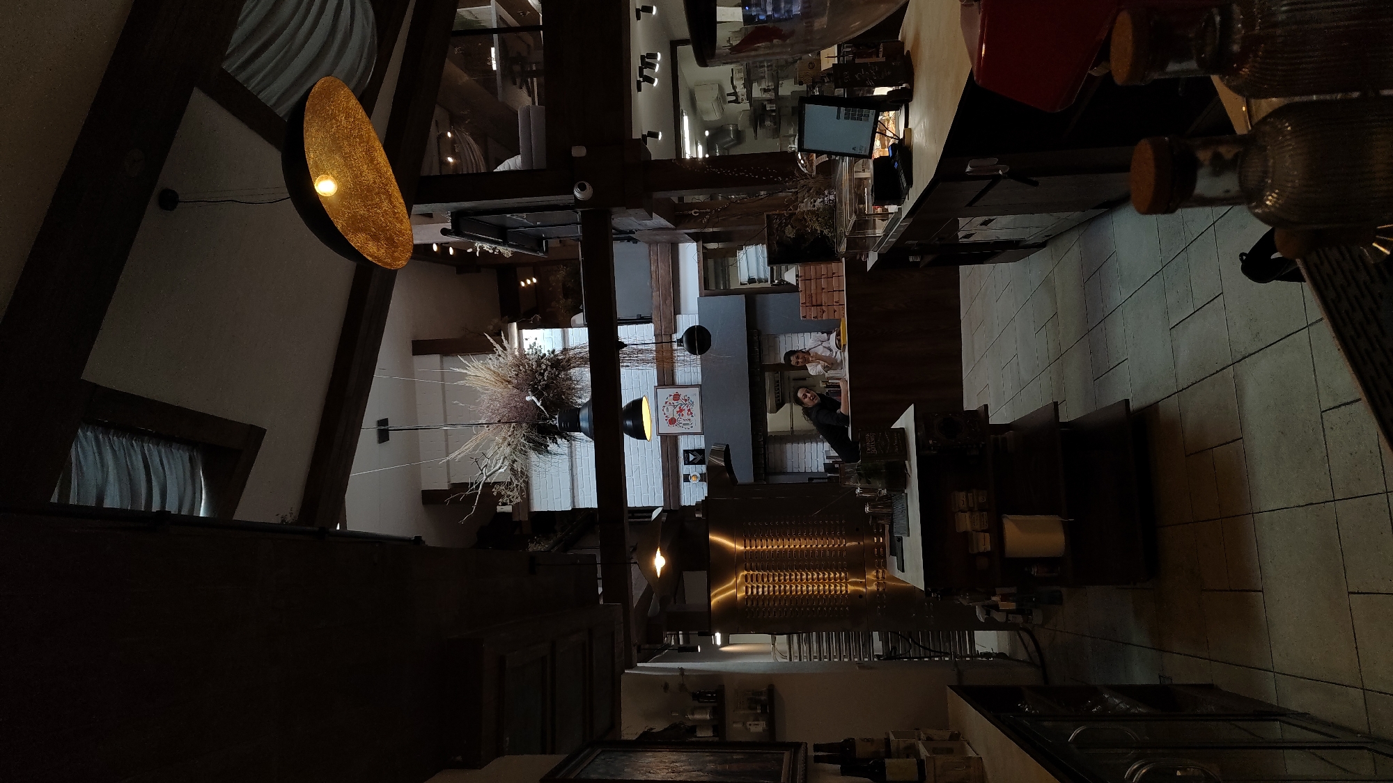 The image depicts the interior of a dimly lit establishment, possibly a cafe or restaurant, with a rustic and industrial aesthetic. Exposed wooden beams traverse the ceiling, with skylights and draped fabric visible. Several pendant lights hang at various heights, one with a prominent golden, textured shade. In the background, two individuals are visible behind a counter. One, with dark hair, leans forward, while the other, wearing a white top, looks towards the first person. A large, stainless steel appliance, resembling an oven or pizza oven, stands prominently to the left, adorned with horizontal ridges. Shelving units with various items are present throughout the space, including bottles on the left and a roll of paper on a shelf in the center. The floor is tiled with a light-colored, grid pattern. The overall impression is one of a cozy, yet modern, dining or service area. Without specific external visual cues, pinpointing the exact location in Plovdiv, Bulgaria, is not possible from this image alone.