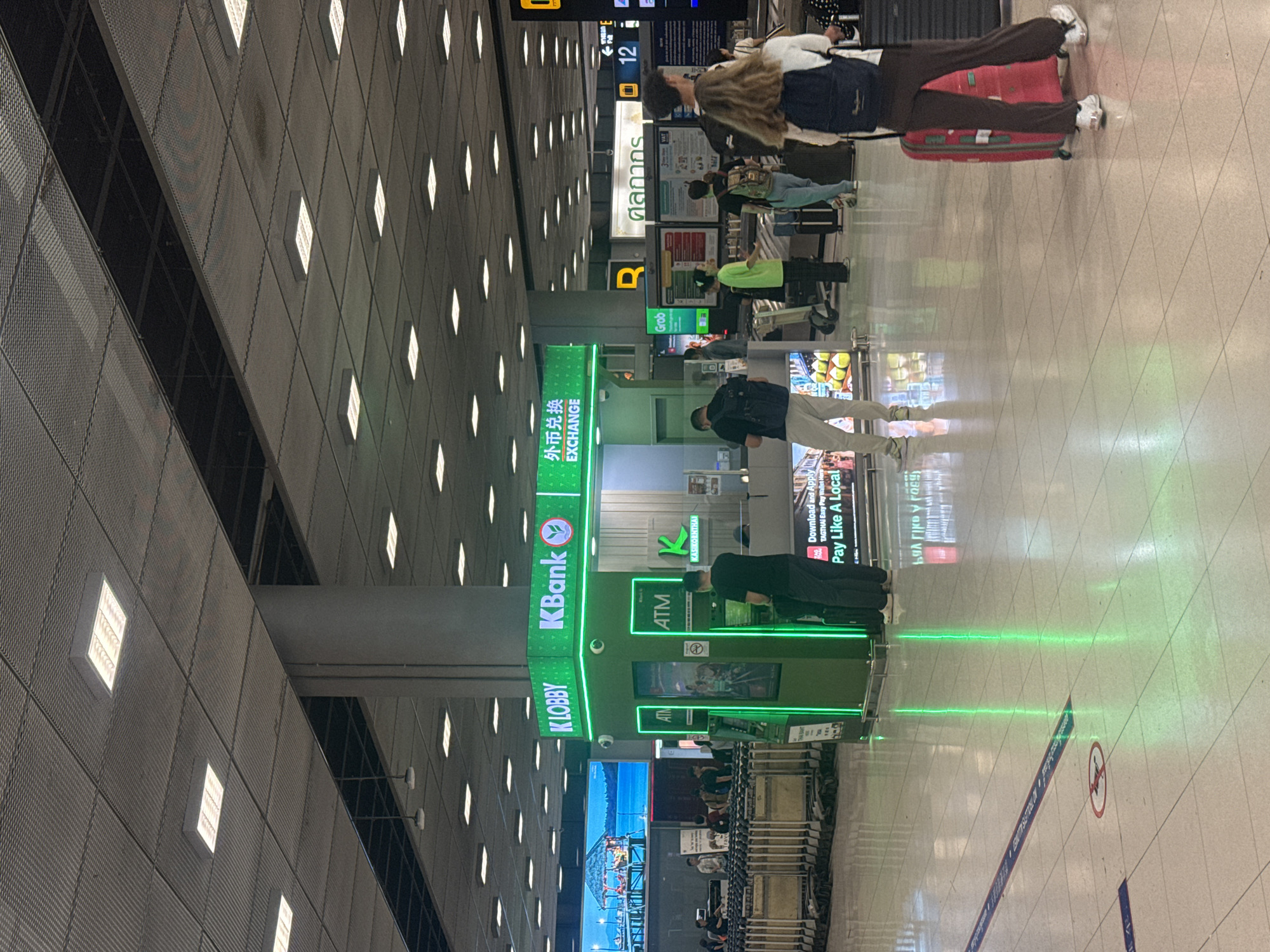 In the city of Nong Prue Subdistrict, Thailand, a busy airport terminal is depicted. A green KBank kiosk is centrally located, featuring ATM machines and foreign currency exchange services. Several people are visible interacting with the ATMs, while others are walking through the terminal with luggage. Shopping carts are lined up to the left of the KBank kiosk, and various signs in Thai and English are displayed throughout the area. The floor is tiled and reflects the bright overhead lighting and the green glow from the KBank kiosk. A large digital screen displaying a tropical scene is visible on the left side of the image.