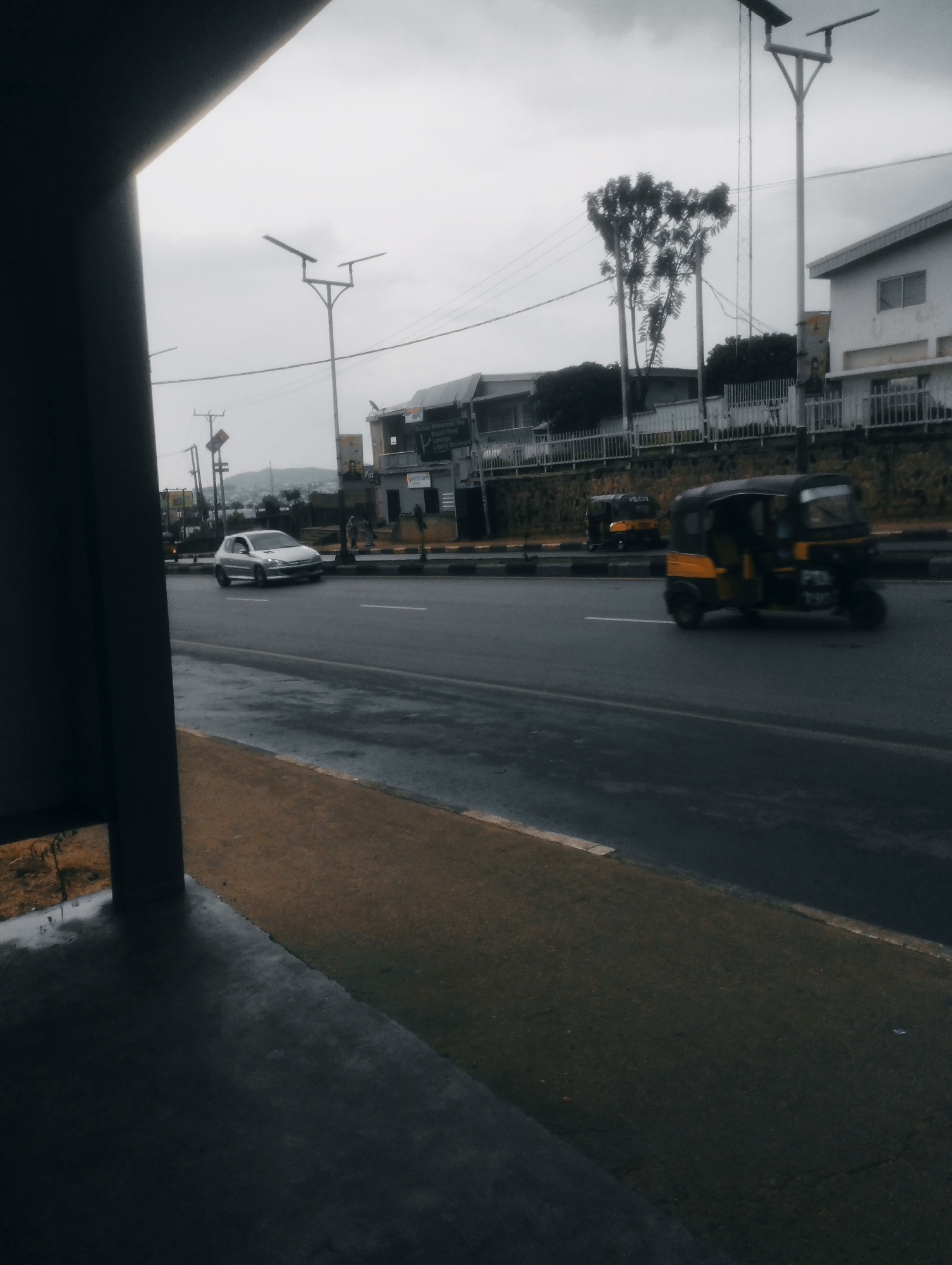 The image depicts a street scene in Jos, Nigeria, on an overcast day. In the foreground, a dark, possibly concrete, covered walkway or bus stop is visible on the left, with its edge extending into the frame. A damp, dark gray asphalt road occupies the middle ground, appearing wet, suggesting recent rain. A yellow and black auto-rickshaw, a common mode of transport in Nigeria, is captured in motion on the right side of the road, its wheels blurred due to movement. Further down the road, another, less distinct auto-rickshaw is visible. To the left of the road, a silver car is driving. Beyond the road, a white building with windows and a fence are present, along with several trees and utility poles. The sky is a uniform gray, contributing to the muted lighting of the scene. The overall impression is that of an ordinary urban environment in Jos, Nigeria, during daylight hours.