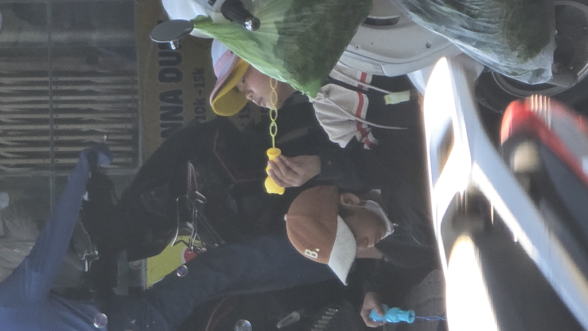 Two individuals are present in the frame. One person, wearing a brown and white baseball cap with the letter "B" on it, is holding a blue bubble bottle. The second person, positioned above the first, is wearing a yellow and pink sun hat and holds a yellow bubble bottle. Bubbles are visible in the lower left of the frame. The location is speculated to be Nha Trang, Vietnam, based on visible text and the context provided. The background includes parts of a vehicle and other unidentified items.