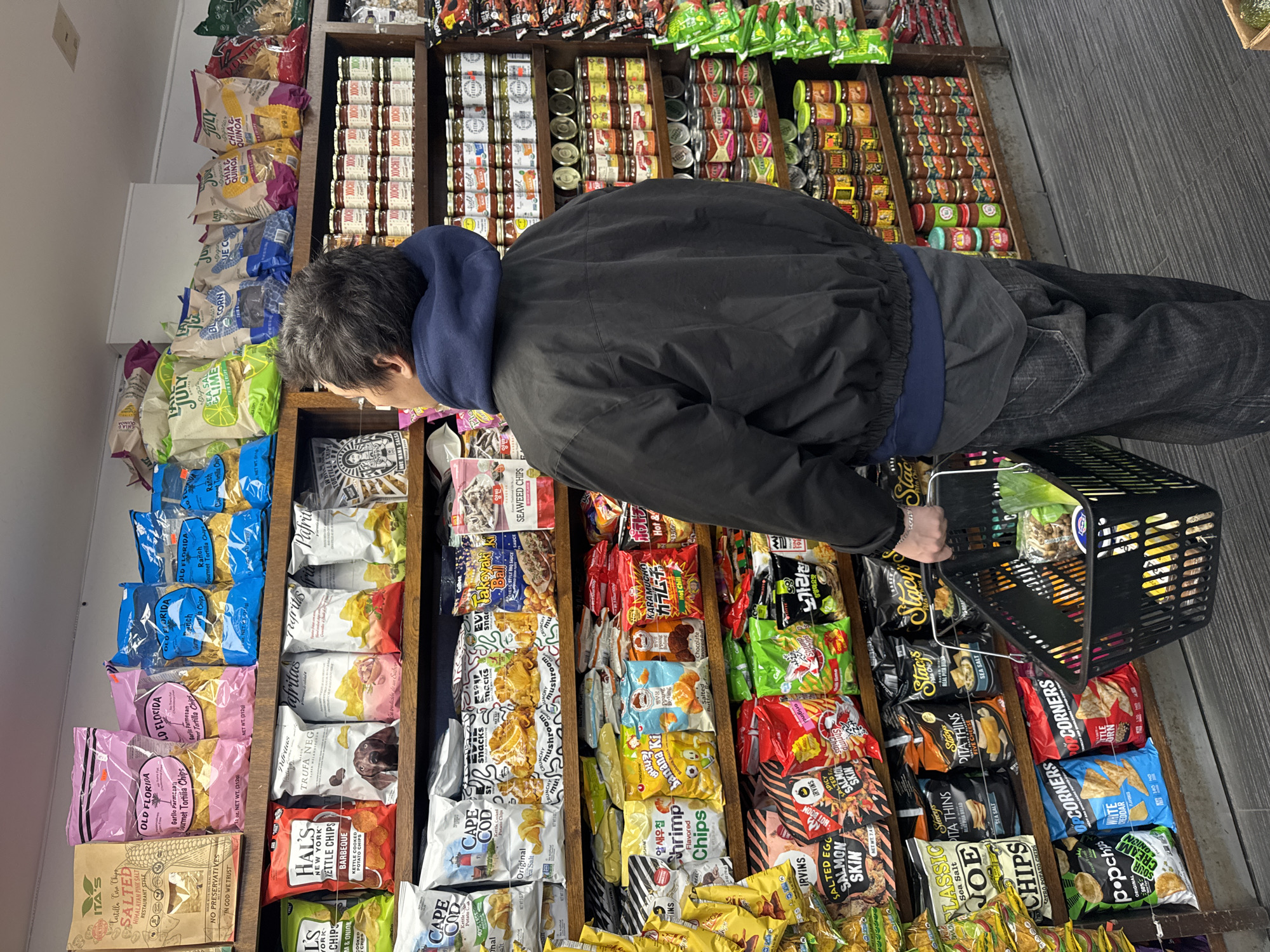 A person wearing a black jacket and jeans is standing in front of a shelf stocked with snacks in what appears to be a store. The person is holding a black shopping basket and looking at the various chip and snack options. The shelves contain a variety of chips, including brands like Old Florida, Cape Cod, Stacy's, and Popcorners, as well as imported snacks. Jars and canned goods are visible on shelves behind the snack chips. The setting suggests a specialty food store or a well-stocked convenience store, possibly in the city of New York based on the Hal's New York Kettle Chips branding.