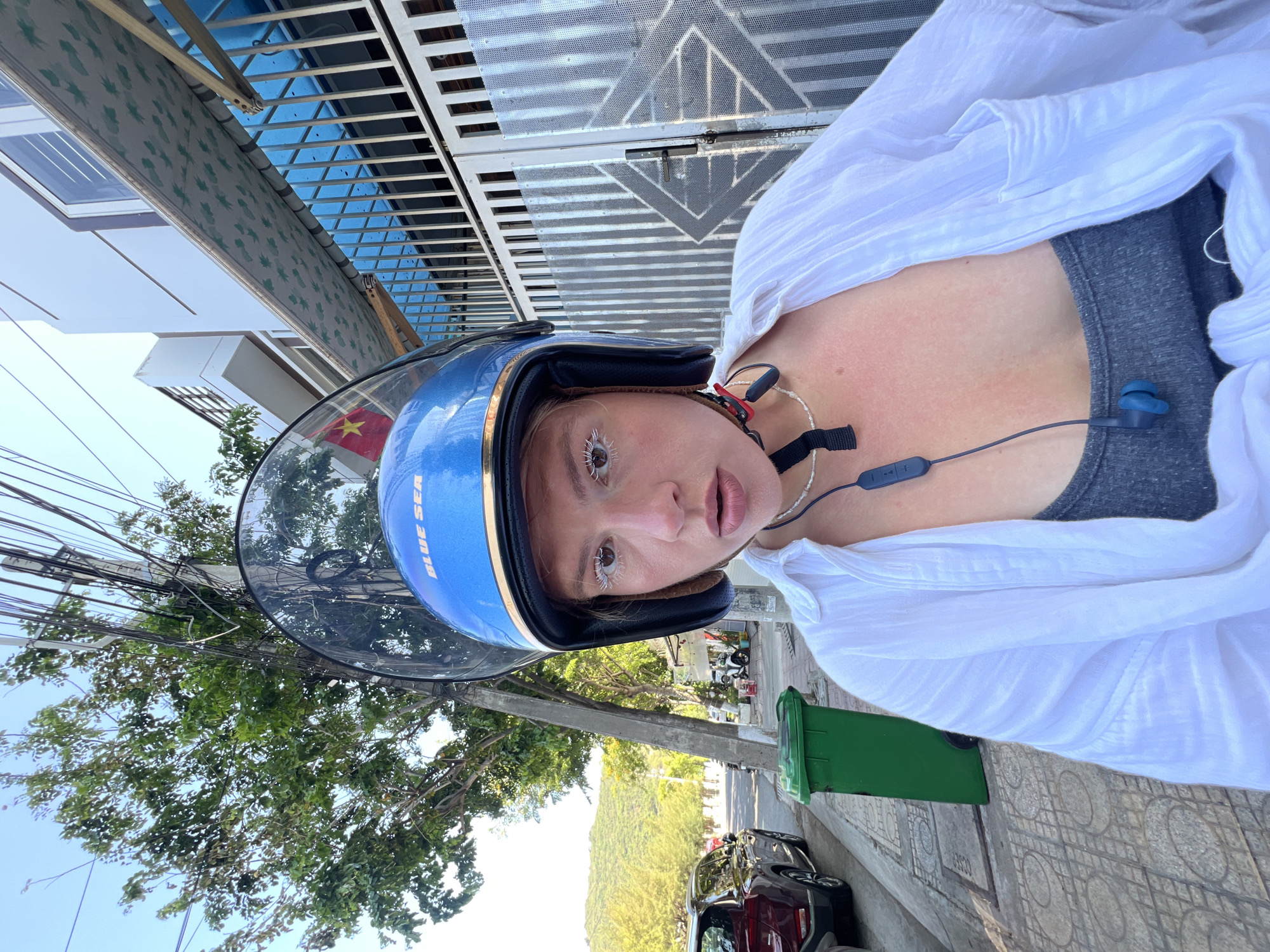A woman wearing a blue motorcycle helmet with the words "BLUE SEA" visible stands on a sidewalk in Nam Nha Trang Ward, Vietnam. She is wearing a gray top under a white shirt and a pair of earphones around her neck. A green trash bin is next to her on the sidewalk. In the background, a street with cars parked along the side leads toward a distant mountain. A Vietnamese flag is visible in the distance. Buildings and foliage line the street. Above, power lines are tangled among the trees. The camera angle is upward, capturing the sky and top of a building with patterned metalwork.