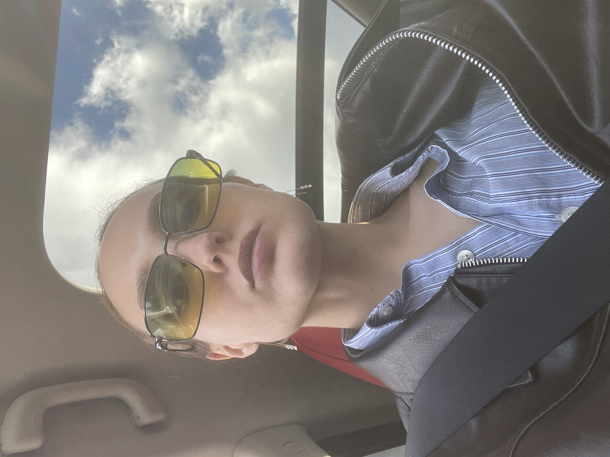 An upward-angled, close-up photograph, originally rotated 90 degrees counter-clockwise, depicts a person inside a car during the daytime.
**Subjects:** The main subject is a person, likely a young adult, with fair skin, wearing yellow-tinted, aviator-style sunglasses. They are dressed in a dark, possibly leather, zippered jacket worn over a light blue and white striped collared shirt. A delicate necklace with small, clear charms is visible around their neck, and an earring is subtly seen on their left ear.
**Activity:** The person is posing for a photo, with their head slightly tilted, and their expression is mostly neutral, partially obscured by the sunglasses.
**Scene and Setting:** The setting is the interior of a vehicle, indicated by a black seatbelt across the lower right, a beige overhead grab handle in the upper left, and the car's interior roof lining. Through a window (likely the windshield or sunroof), a bright blue sky with scattered white clouds is prominent, suggesting a clear day. The natural light illuminates the person and the car interior.
**Notable Details:** The jacket features a distinct silver zipper. The striped shirt has fine blue and white lines. The weather is sunny with some cloud cover. No discernible text is visible in the image. The location context for this scene is Ol'khovochka, Russia.