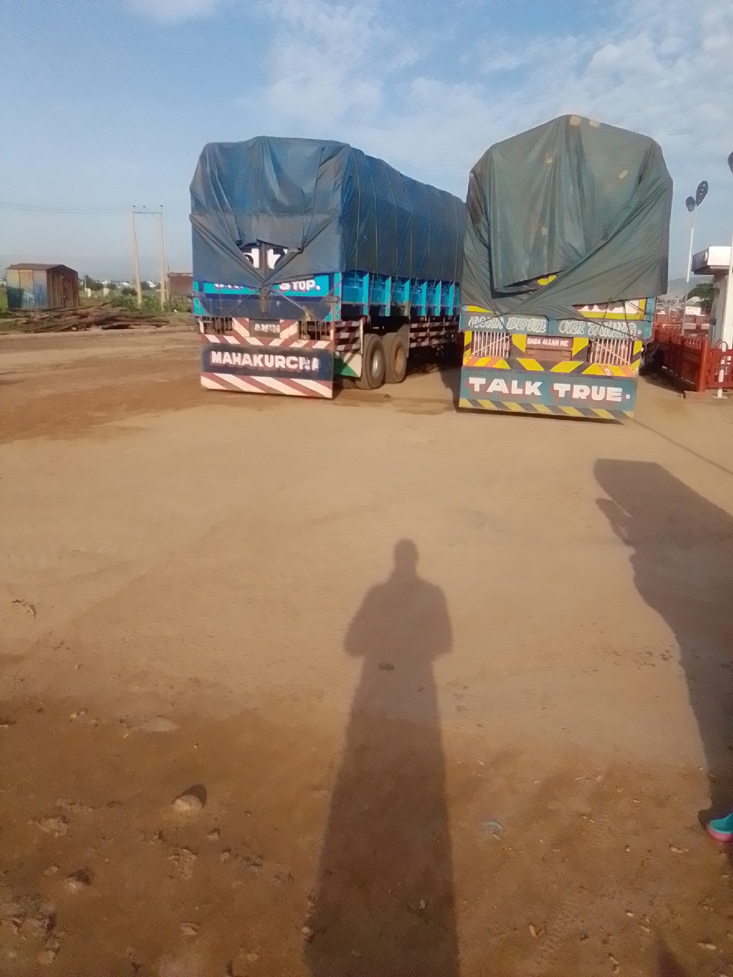 Two large trucks are parked side-by-side on a dirt lot, likely in Kwadon, Nigeria. The truck on the left has a blue tarpaulin covering its cargo, and the truck on the right has a dark green tarpaulin. Both trucks have colorful markings on their rear ends. The truck on the left displays "MAHAKURCHI" in white letters on a blue and red striped background. The truck on the right has "TALK TRUE" written in large white letters on a teal background, with yellow and black striped accents above. The ground is dusty and uneven, with scattered small stones and debris. In the background, there are some wooden logs, a small building with a corrugated metal exterior, and power lines. The sky is partly cloudy with a bright blue hue. A shadow of a person, possibly the photographer, is cast on the ground in the foreground. The scene appears to be an open area, possibly a transport hub or a roadside rest stop.