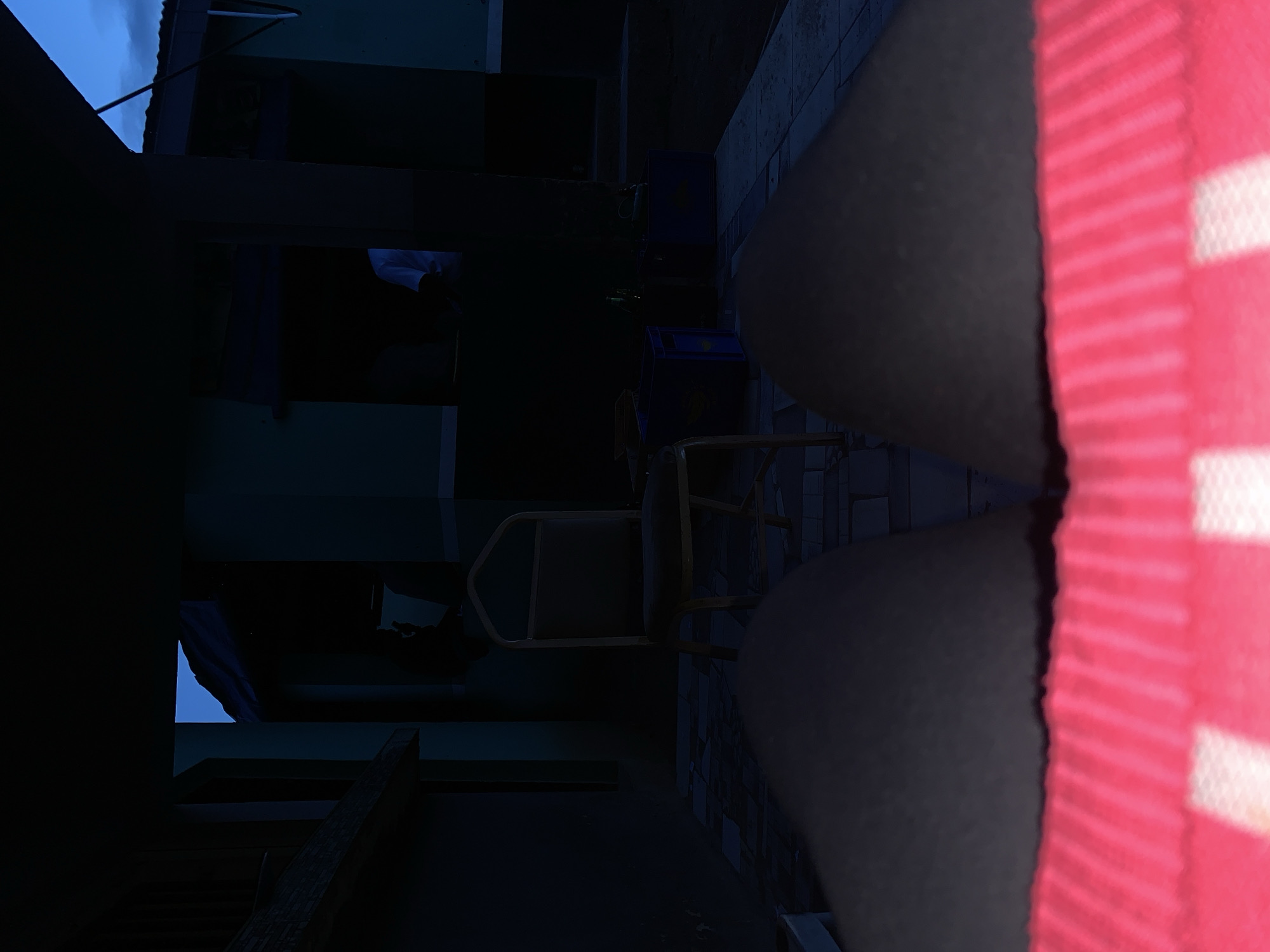 The image, captured in New Stadium, Nigeria, presents a low-light outdoor scene from a low-angle perspective. In the immediate foreground, the upper thighs of an individual are visible, wearing clothing characterized by red and white horizontal stripes. The ground surface in the midground consists of dark-toned tiles. An empty chair with a metallic frame and dark seat is positioned on the tiled surface, facing away from the camera. To the right of the chair, two blue plastic crates and several dark-colored bottles are present on the ground. In the background, a person wearing a white T-shirt is partially visible through an opening within a dimly lit building structure. The surrounding environment comprises multiple dark-walled buildings, structural pillars, and rooflines. The sky, visible through openings between structures, is a deep blue, suggesting either twilight or pre-dawn conditions. The scene is illuminated solely by ambient natural light.