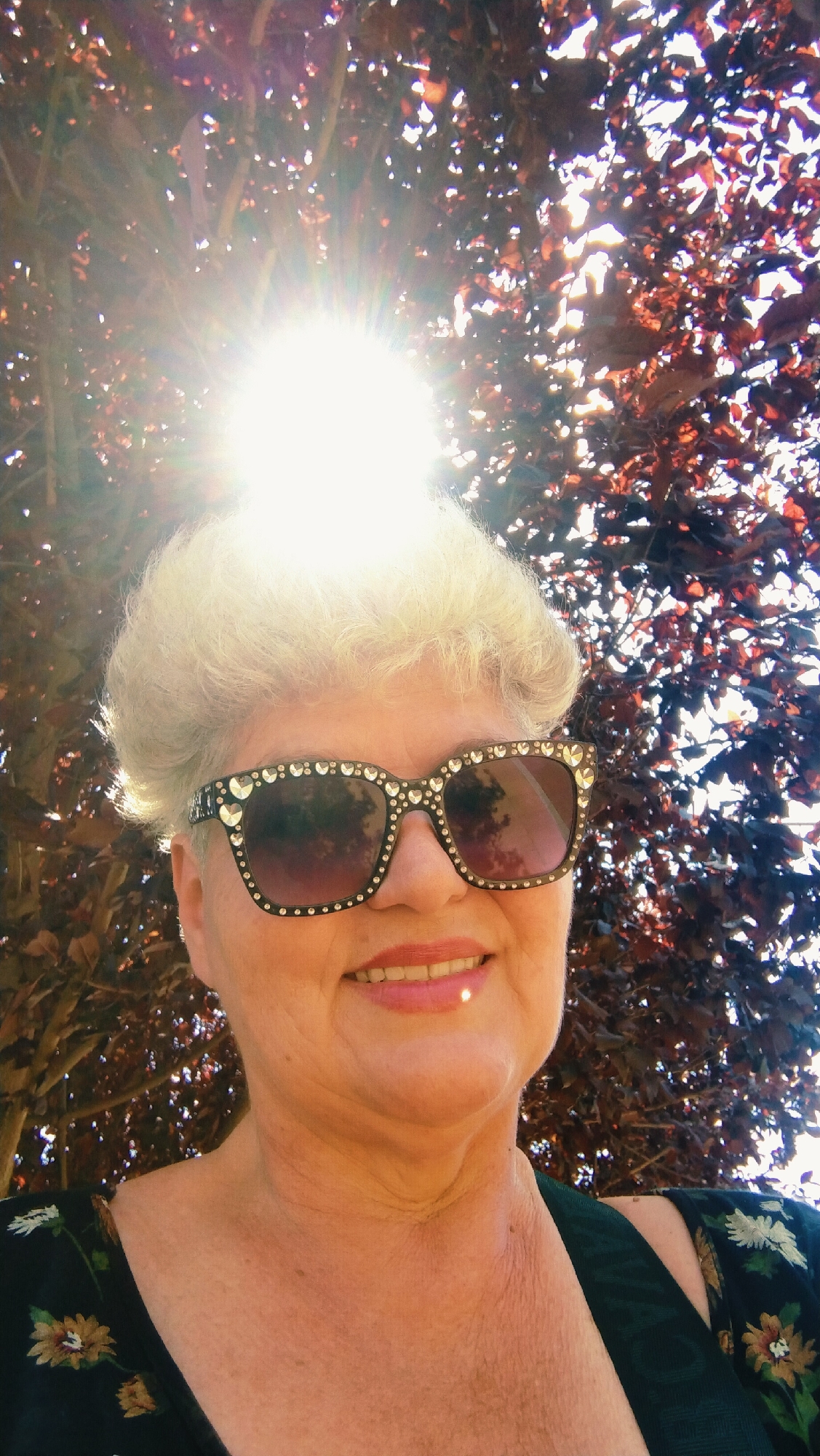 A close-up, waist-up, outdoor selfie shows a fair-skinned, middle-aged woman with blonde, styled hair, wearing large, black sunglasses with rhinestones and heart-shaped accents, and a dark floral print top. The woman is smiling and her lips are painted with pink lipstick. She is positioned in front of a tree with dark red leaves, with the sun prominently visible behind and to the left of her head, creating a bright lens flare effect. The background foliage is out of focus. The image was taken at eye level. The woman is looking directly at the camera. The image was taken in Horishni Plavni, Ukraine.