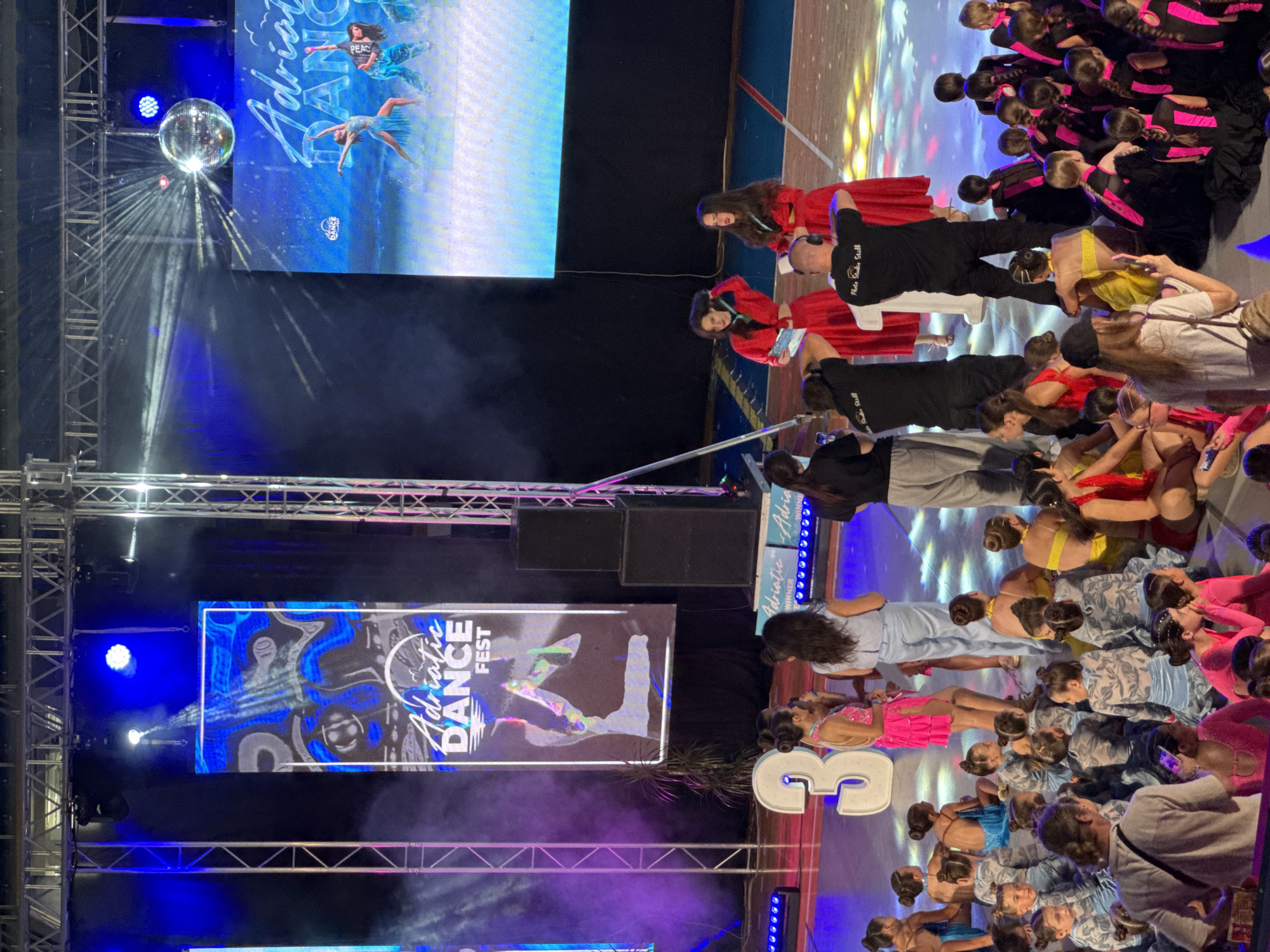 The image captures a dance competition event taking place indoors, possibly in a sports hall or convention center in Bar, Montenegro. In the background, large screens display the "Adriatic Dance Fest" logo and dynamic imagery of dancers. A prominent "3" shaped prop is visible on the stage, alongside a group of young female dancers in various colorful costumes. Some dancers are seated on the stage floor, while others are standing. Two women in red dresses are on a raised platform, possibly receiving awards, with a photographer in the foreground. The lighting is dramatic, with spotlights and colored lights illuminating the stage and creating a festive atmosphere. A disco ball hangs from the ceiling, reflecting light. The overall scene suggests an energetic and celebratory dance event.