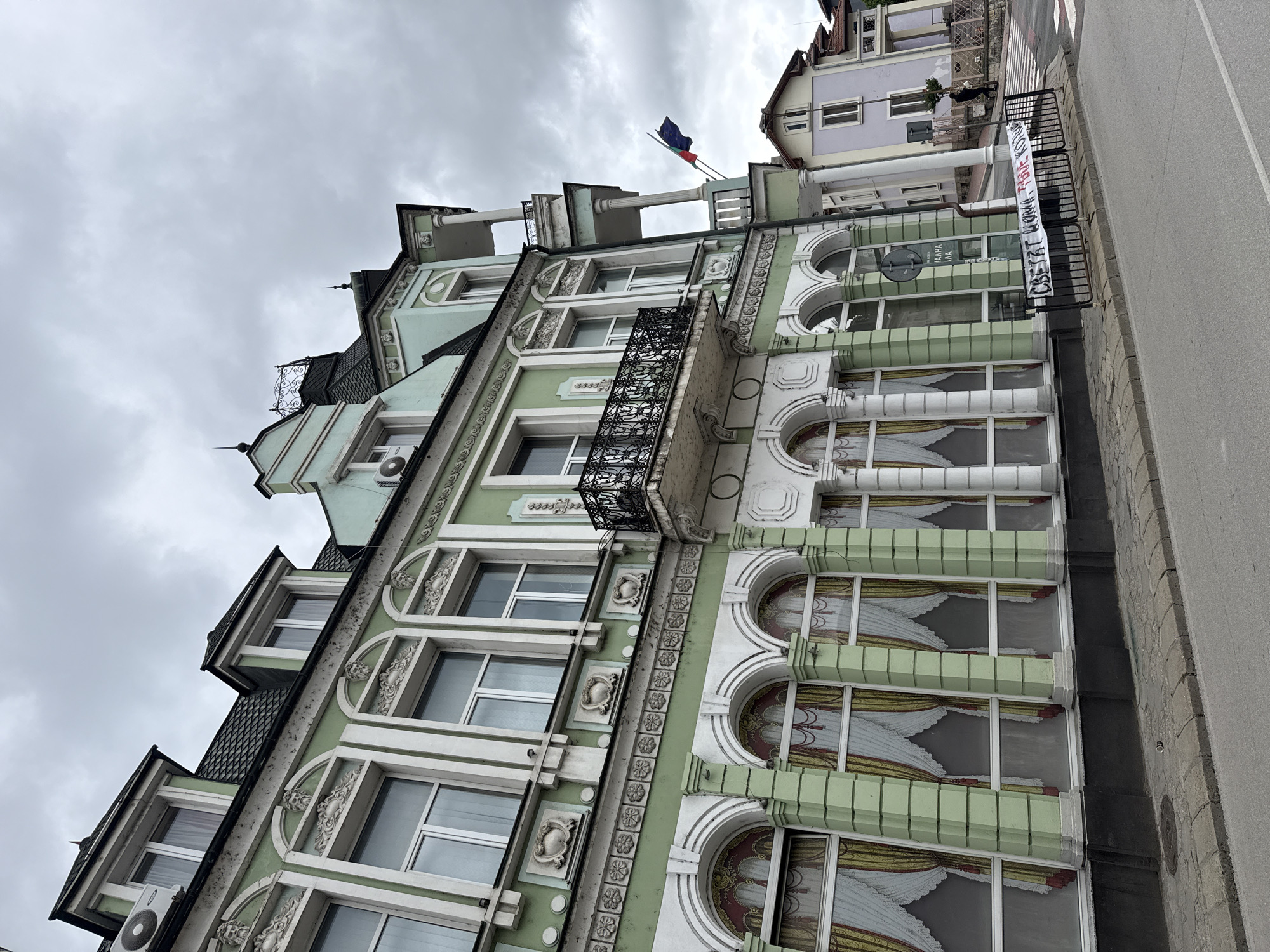 The image depicts a multi-story building in Lovech, Bulgaria. The building has a light green facade with white architectural details. The windows on the lower floors have arched tops with ornate decorations. A small balcony with black metal railings is visible on one of the upper floors. A banner with Cyrillic script is attached to the railing of an adjacent building. Two flags, the Bulgarian and European Union flags, are seen on the roof of a neighboring building. The sky is overcast.