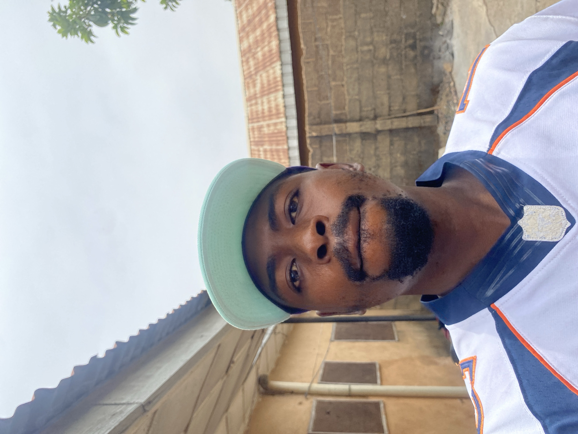 An upward-angled, close-up shot captures a man with dark skin, a beard, and a mustache, looking directly at the camera. He wears a light blue or mint green baseball cap, reversed, and a white sports jersey with prominent blue and orange stripes. The number "17" is partially visible on the right sleeve, and a faded patch or logo is on the right shoulder. His expression is neutral.
The background consists of an overcast, light grey sky dominating the upper left and center. Green leaves from a tree are visible in the top left corner. Weathered buildings form the rest of the background. To the right, there is a reddish-brown brick wall. Further back and to the left, a structure with a corrugated metal roof is visible, and below it, a lighter-colored building, possibly yellow or beige, featuring multiple dark, rectangular openings and an exterior pipe. The lighting suggests it is daytime, possibly under cloudy skies. The architectural elements are consistent with the location context of Barakin Akawo, Nigeria.