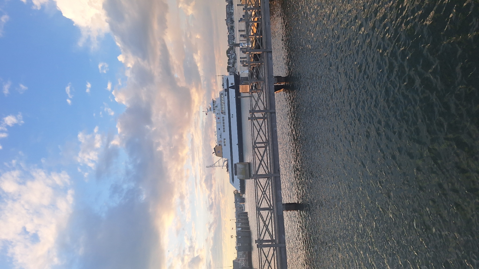 The image depicts a ferry docked at a harbor in Nantucket, United States of America. The ferry, a large white vessel with a yellow stripe and black trim, is positioned in the background. In the foreground, a wooden pier with a crisscross railing extends into the dark, rippling water. The sky above is a mix of blue and clouds, with warm tones suggesting either sunrise or sunset. A few other boats are visible in the distance, as well as buildings along the coastline. The scene is calm and serene, with no people visible.
