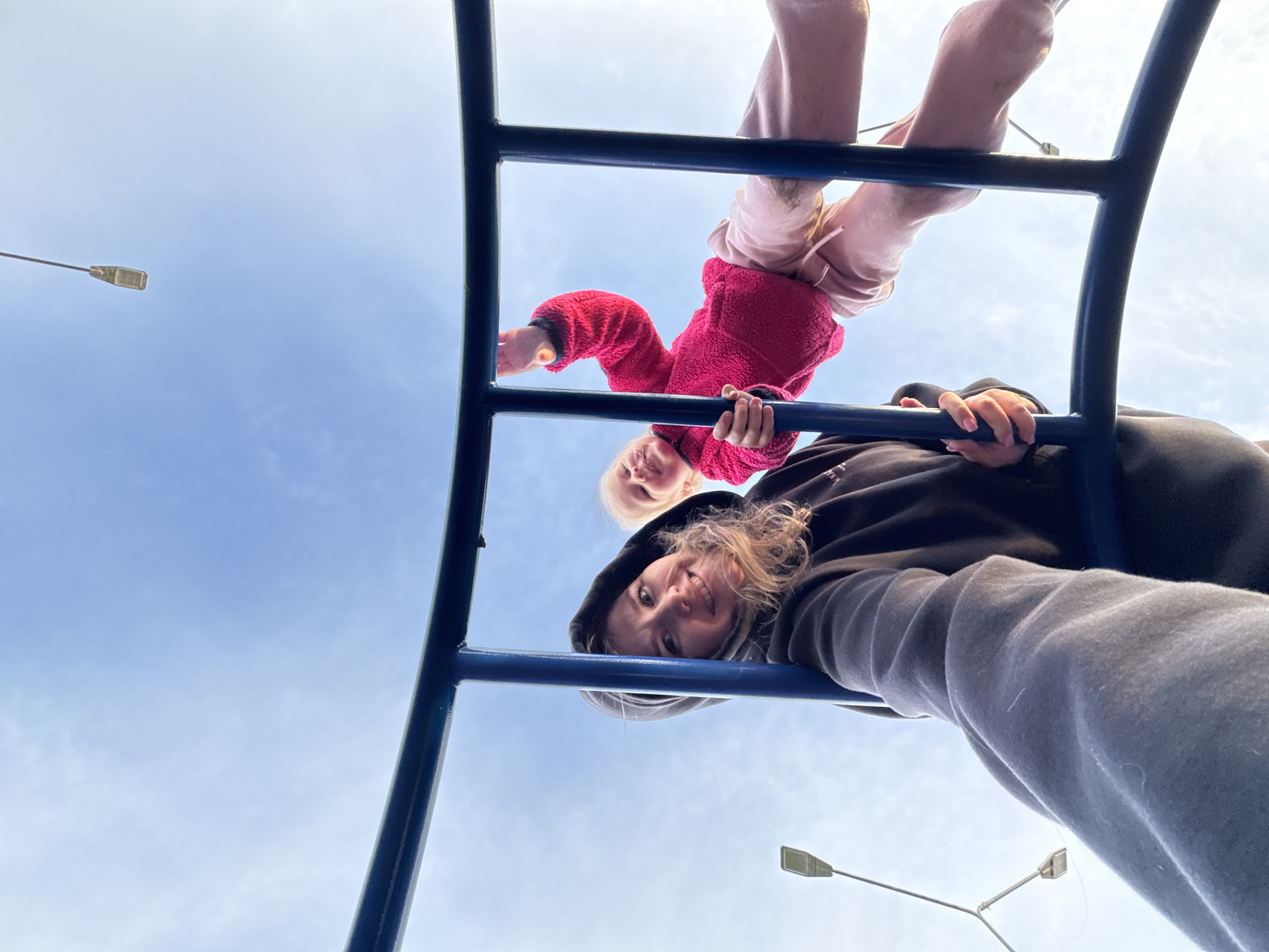 The image displays an adult female and a young child interacting with a blue metal playground structure, viewed from a low-angle perspective against a bright, clear blue sky with minimal cloud cover. The scene is located in the city of Горный, Russia.
In the left foreground, an adult female with light-colored hair, wearing a dark hooded sweatshirt, is positioned with her face visible and directed towards the camera, exhibiting a smile. Her right arm extends downwards, gripping a vertical blue bar of the structure.
To the right and slightly above the adult, a young child with blonde hair is situated within the blue structure. The child is clad in a bright pink fleece top and light pink pants. The child's face is visible, smiling and oriented towards the camera. The child's left hand is raised in a waving gesture, while the right hand grasps a vertical blue bar. The child's lower body and legs are visible, positioned across the horizontal bars.
Both individuals are engaged with the blue metal bars, suggesting climbing, hanging, or posing within the structure. The low camera angle indicates a close-up shot, potentially a selfie.
In the upper and left background, multiple modern streetlights on tall poles are visible against the sky. The lighting conditions suggest an outdoor daytime setting with fair weather.