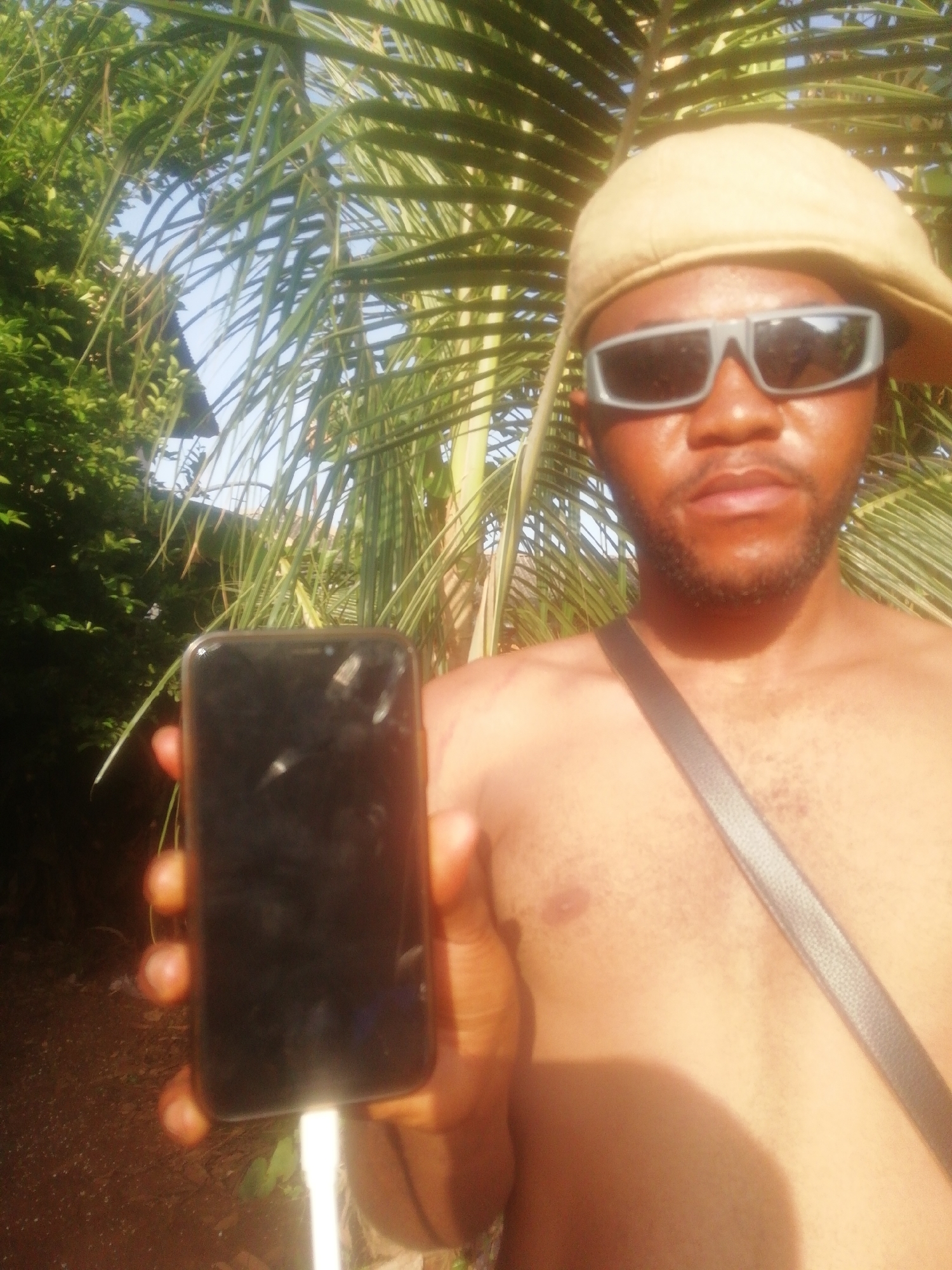 The image depicts a dark-skinned man standing outdoors. He is shirtless, wearing a tan baseball cap and sunglasses. A black strap crosses his chest, presumably from a bag. He is holding a black smartphone, which is plugged into a white charging cable. The background features lush green vegetation, including palm fronds, suggesting a tropical environment. The location is listed as Unknown, Unknown.