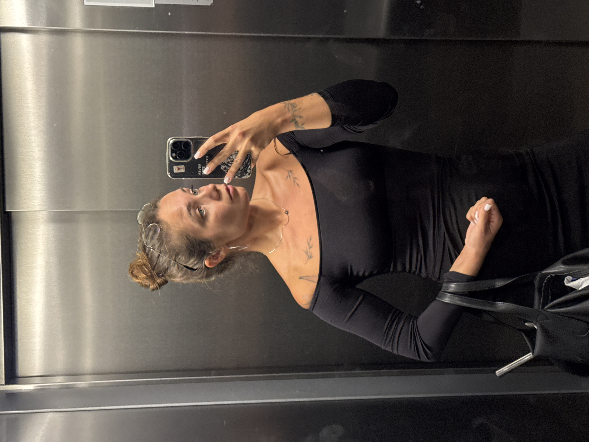 A woman is taking a mirror selfie in an elevator. She has her hair pulled up in a bun and is wearing a black off-the-shoulder dress. She has a thin necklace on and a small tattoo on her shoulder. She is holding a black purse in her left hand. The elevator walls are made of stainless steel. The photo appears to have been taken in Budva, Montenegro.