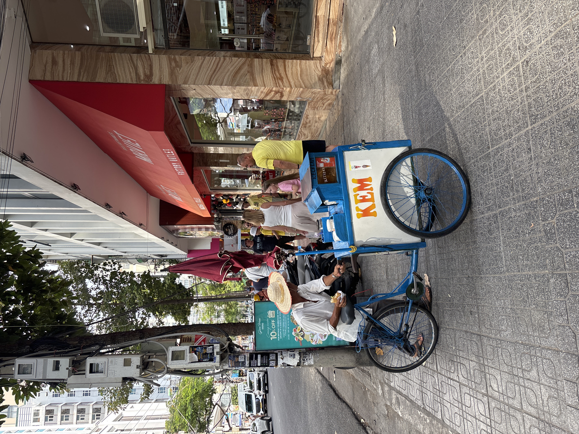 The image depicts a street scene in Nha Trang Ward, Vietnam. A vendor wearing a straw hat and a white shirt is seated on a blue bicycle attached to a mobile ice cream cart. The cart is white with blue trim and has "KEM" (meaning ice cream in Vietnamese) written in large orange letters on its side. The vendor is holding a piece of food, possibly an ice cream cone, and appears to be eating. The cart also displays an advertisement for "Green Home Dining" offering a "10% OFF" discount.
The background shows a sidewalk paved with grey patterned tiles. To the right of the sidewalk, there is a building with a red awning and large glass windows, displaying merchandise inside. Several people are walking on the sidewalk, including a woman in a white top and light-colored pants, a young girl in a pink shirt, and a man in a yellow t-shirt and black shorts. A red umbrella is also visible on the sidewalk, and several motorcycles are parked along the street.
The street itself is paved with asphalt and has traffic, including white vans and cars. A traffic light pole with several electrical meters is located to the left of the vendor. Trees with green foliage are present along the sidewalk and street. The overall atmosphere is that of a bustling urban environment on a sunny day.