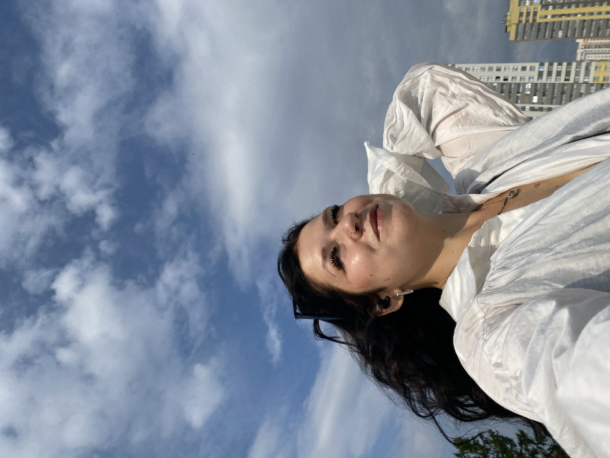 A young woman with long, dark hair stands outdoors, looking up at a cloudy blue sky. She is wearing a white, voluminous shirt with puffed sleeves. A silver necklace with a pendant is visible at her décolletage. She has a nose ring and earrings. In the background, two tall, modern buildings are visible. The woman is holding one hand behind her head, her arm bent. The overall impression is of a casual, possibly windy day in an urban environment.