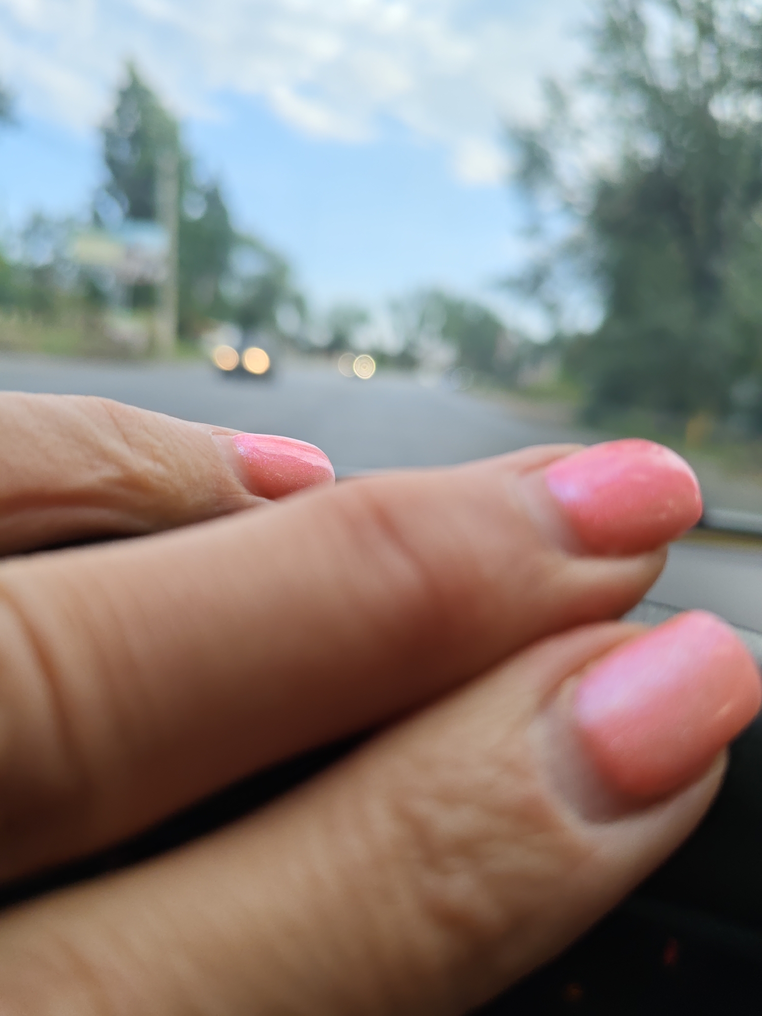 In Novocherkassk, Russia, a close-up shot focuses on a person's hand with pink nail polish, positioned in the foreground and blurring into the background. The background shows a street scene with out-of-focus cars and trees under a cloudy sky. The overall impression is a snapshot taken from inside a vehicle.