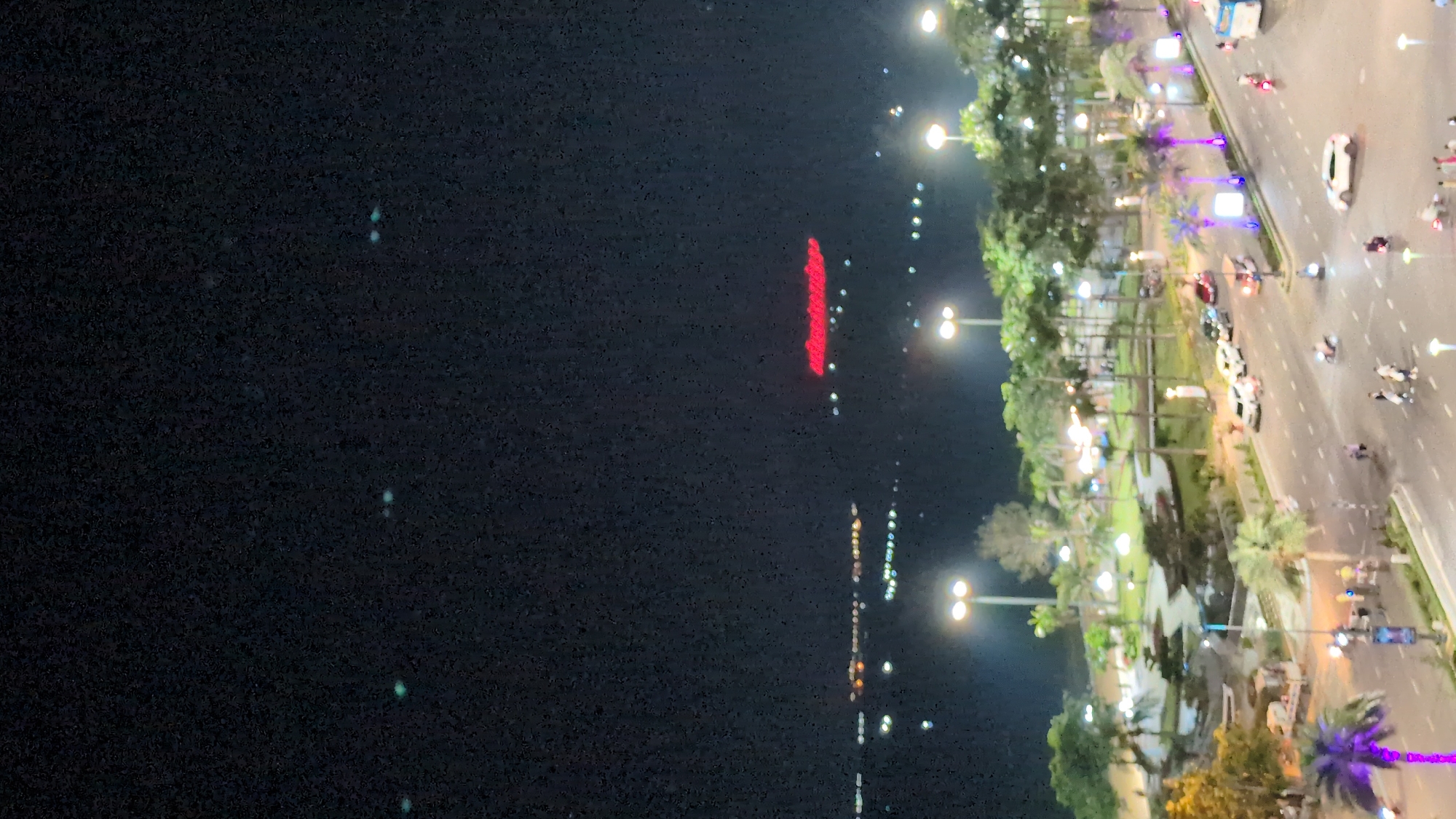 This nighttime image presents an elevated view of a coastal urban landscape in South Quy Nhon Ward, Vietnam. The upper portion of the frame is dominated by a dark sky, exhibiting several faint, greenish-blue light anomalies.
The midground features a brightly illuminated multi-lane boulevard running horizontally, lined with white demarcation lines. Numerous vehicles are present on the road, including white, red, and dark-colored cars, several motorbikes in motion, a white SUV, and a partially visible blue bus or large vehicle on the right. A white utility truck is observed on the left side of the road. Multiple pedestrians are visible on the sidewalks and near crosswalks, engaged in walking.
Adjacent to the boulevard, a landscaped park or green space extends towards the water, characterized by numerous palm trees and other dense foliage, all brightly lit by streetlights. Some palm trees display decorative purple illumination. The park features winding pathways and additional ambient lighting.
In the background, a dark body of water, likely a bay or ocean, stretches into the distance. It is dotted with numerous bright, indistinct lights, indicative of distant vessels or structures along a remote shoreline. Prominently positioned above the water in the far distance is a large, horizontally elongated red illuminated sign, which appears to spell "FLC LUXURY".