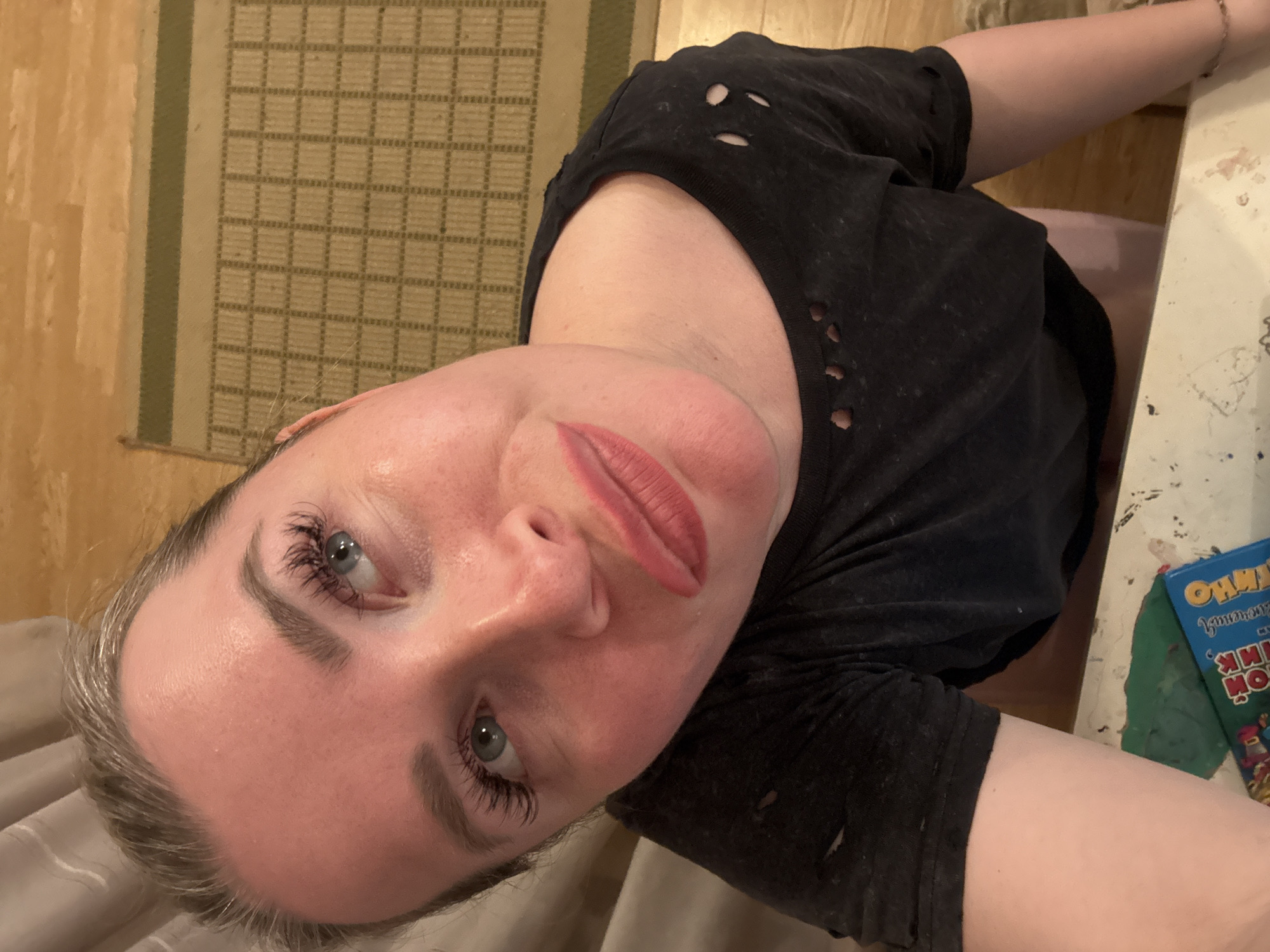 A Caucasian woman, likely in her 20s or 30s, is captured in a close-up, high-angle selfie in Veliky Novgorod, Russia. Her light-colored eyes (blue/grey) are emphasized by prominent eyelashes and groomed eyebrows, and her light-colored hair is styled back. She wears pinkish-red lipstick on slightly parted lips, displaying a subtle smirk. Her attire consists of a black, distressed t-shirt with visible holes around the collar and shoulders. The camera is positioned low, angled upwards, creating a wide-angle perspective that emphasizes her head. In the immediate foreground, a light-colored surface, possibly a table, shows paint splatters or marks. On this surface, a blue rectangular object, resembling a board game box with colorful graphics and partial text like "CAMO," is visible next to a blob of green material. The background includes light-colored curtains on the left and wooden floorboards on the right, partially covered by a beige-and-brown woven mat with a grid pattern.