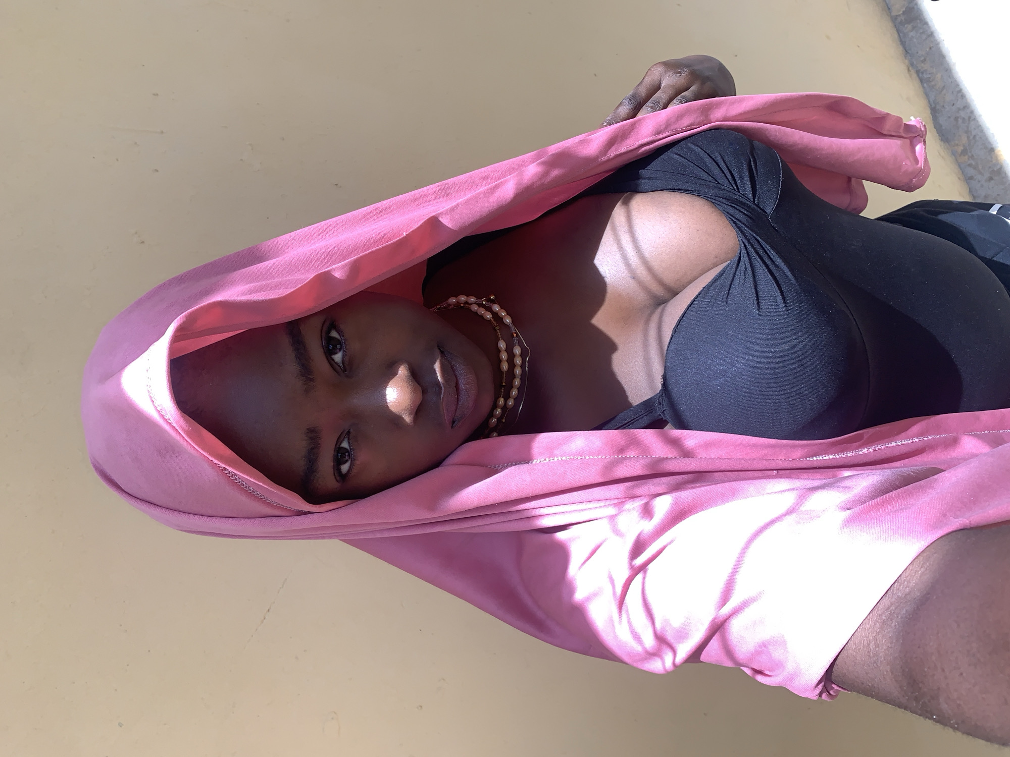 A woman with dark skin is wearing a black top and a pink headscarf. She is also wearing a pearl necklace. The background is a light yellow wall. The photo appears to be taken in an outdoor setting. There is no specific indication of location within the city of Rafin Par, Nigeria.