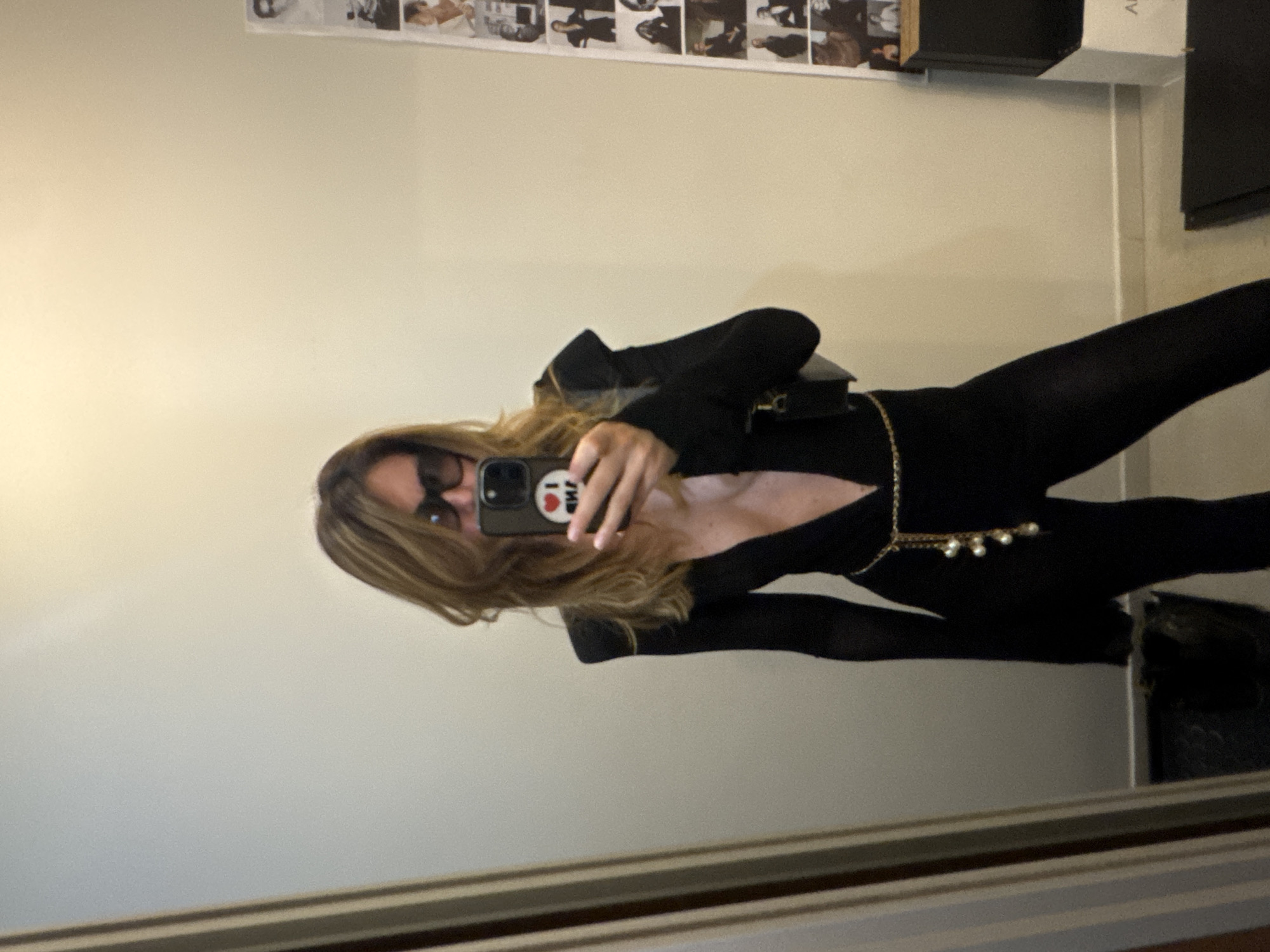 A person is taking a mirror selfie in what appears to be a room in Sofia, Bulgaria. The person is wearing a black jumpsuit with a deep V-neck, sheer sleeves, and a gold chain belt with pearl embellishments. They are also wearing dark sunglasses and holding a smartphone. The background shows a wall with a collage of black and white photographs, a large mirror, and some furniture. The lighting is warm and diffused.
