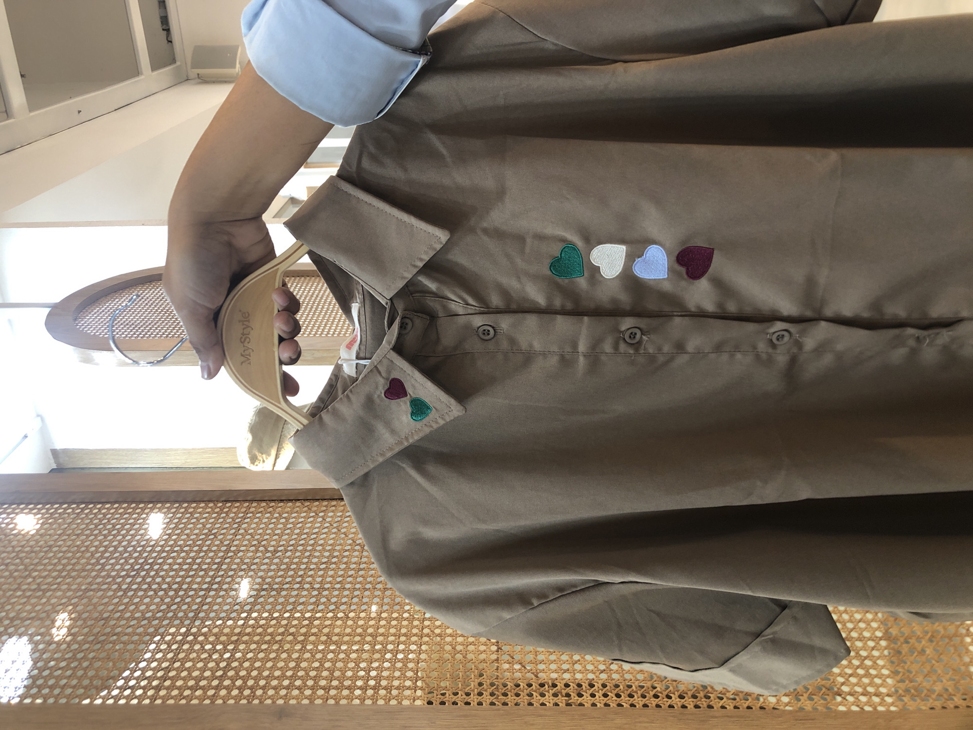 A close-up, eye-level shot shows a light brown collared shirt with embroidered hearts hanging on a wooden hanger. The hearts, in colors of burgundy, green, white, and light blue, are arranged vertically down the front of the shirt. The shirt has short sleeves and visible buttons. A hand holding the hanger is visible, wearing a light blue long-sleeved shirt. In the background, a wooden partition with a woven pattern and some bright light reflections can be seen. The scene appears to be indoors, possibly in a retail store. The image was taken in Surabaya, Indonesia.