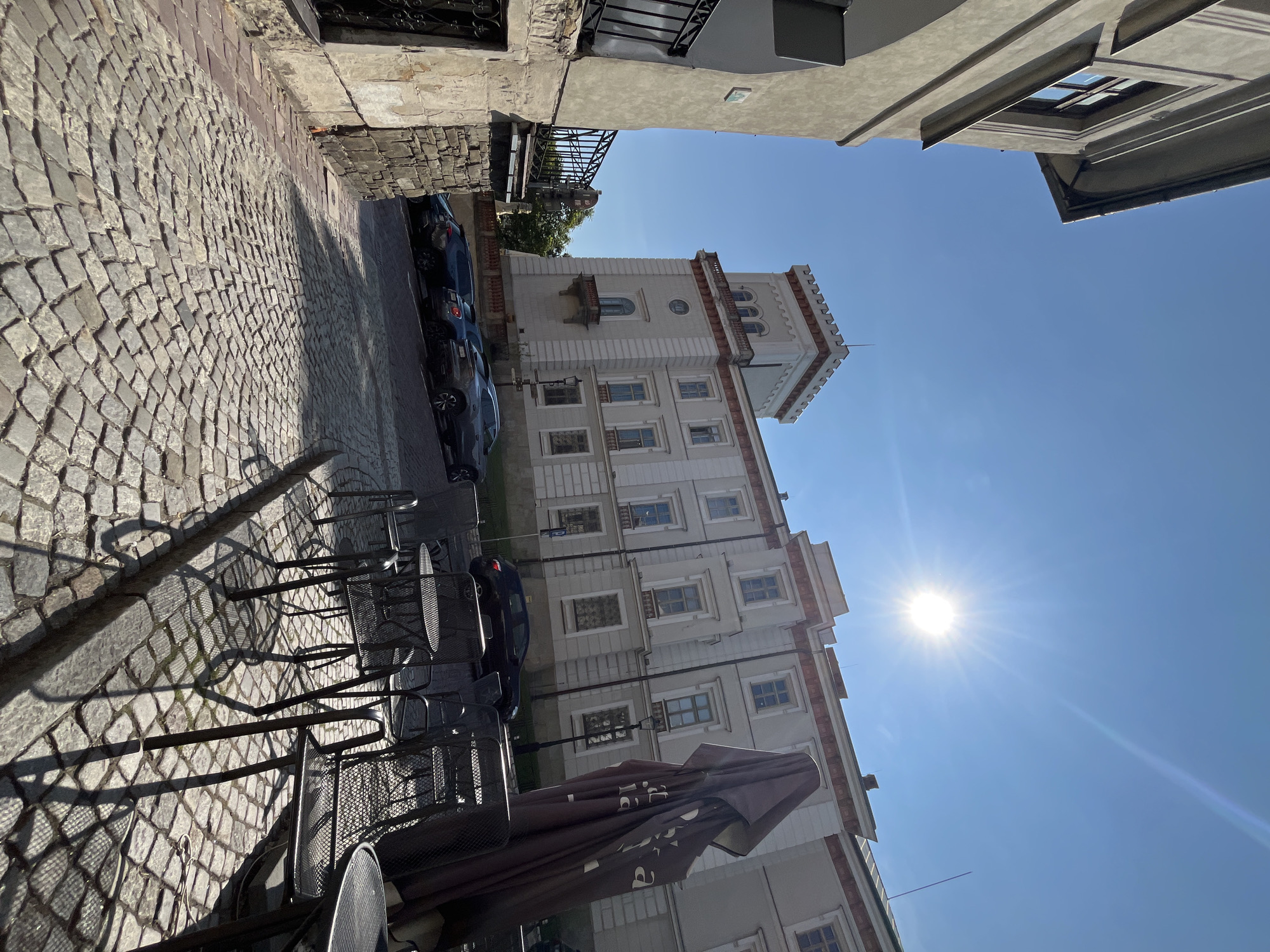 This is a low-angle, wide shot of a cobblestone street in front of a light-colored, multi-story building with ornate architectural details, including a prominent tower with a crenellated top. The building has multiple windows with dark frames and a balcony extending from the tower. Several cars are parked along the street, which curves to the right. In the foreground, there are several wrought-iron tables and chairs set up, with a large, dark umbrella in the process of being closed. The sun is bright and high in a clear blue sky, casting long shadows across the scene. The building appears to be a historic structure, possibly a museum or administrative building, situated in an urban environment.