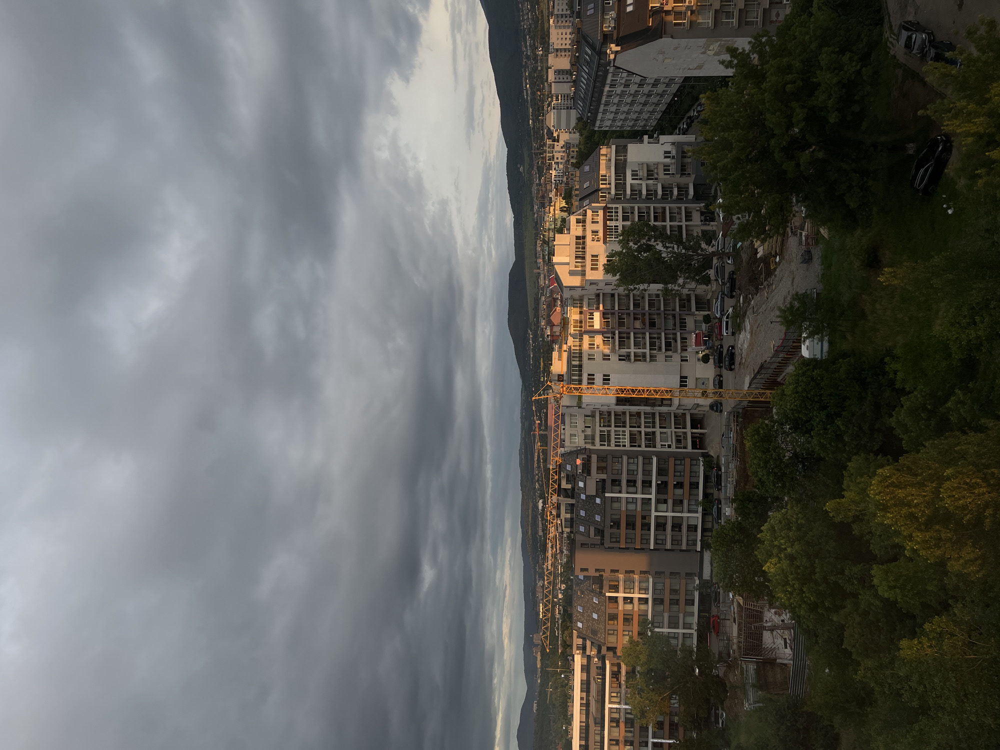 An aerial, wide shot captures a city skyline under a cloudy, overcast sky. In the foreground, a construction site with wooden scaffolding and a white trailer is visible amongst lush green trees. The midground features several modern apartment buildings, some illuminated by the setting sun, and a tall yellow construction crane. Further in the distance, more buildings and cranes dot the landscape, with rolling green hills forming the background. Several cars are parked on the street and in parking areas.