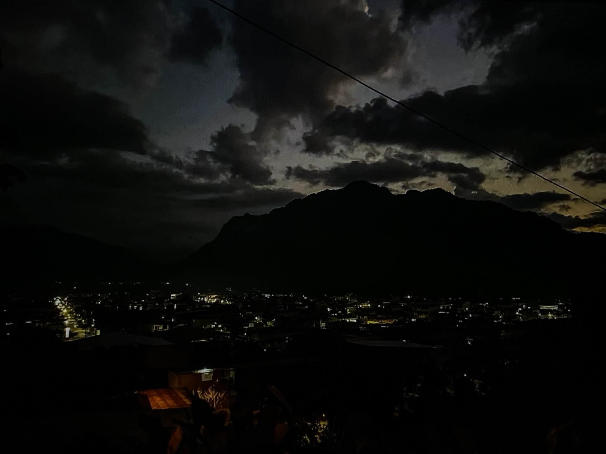The image captures a nocturnal landscape showcasing the city of Tinaquillo, Venezuela, nestled against the silhouette of imposing mountains under a cloudy sky. The scene is dominated by deep blues, grays, and blacks, characteristic of nighttime, with artificial lights illuminating the urban sprawl.
In the upper two-thirds of the frame, a dark, heavy cloud cover stretches across the sky. Patches of lighter, yellowish-grey hues are visible on the horizon, hinting at either the last remnants of dusk or the faint glow of an approaching dawn, possibly reflecting distant light sources or a sliver of natural light. A prominent, dark utility line or cable diagonally traverses the upper right portion of the sky, adding a man-made element to the natural vastness.
Below the expansive sky, the colossal silhouettes of mountains rise, their jagged peaks forming a stark, dark barrier against the clouds. They appear as a formidable backdrop to the human settlement.
At the base of these mountains, the city of Tinaquillo sprawls, a network of twinkling artificial lights. These scattered points of light, predominantly yellow and white, outline streets and buildings, indicating a vibrant urban area. The density of lights suggests a populated cityscape extending into the mid-ground.
In the immediate foreground, particularly towards the bottom left, a dimly lit area reveals what appears to be part of an outdoor setting, possibly a patio or balcony. A wooden surface, perhaps a table, is faintly visible, along with dark, indistinct shapes that could be furniture or figures, mostly obscured by the deep shadow.
The overall mood is serene and contemplative, highlighting the contrast between the dark, natural elements of the mountains and sky, and the glowing, active human presence in the valley. There is no visible text in the image.