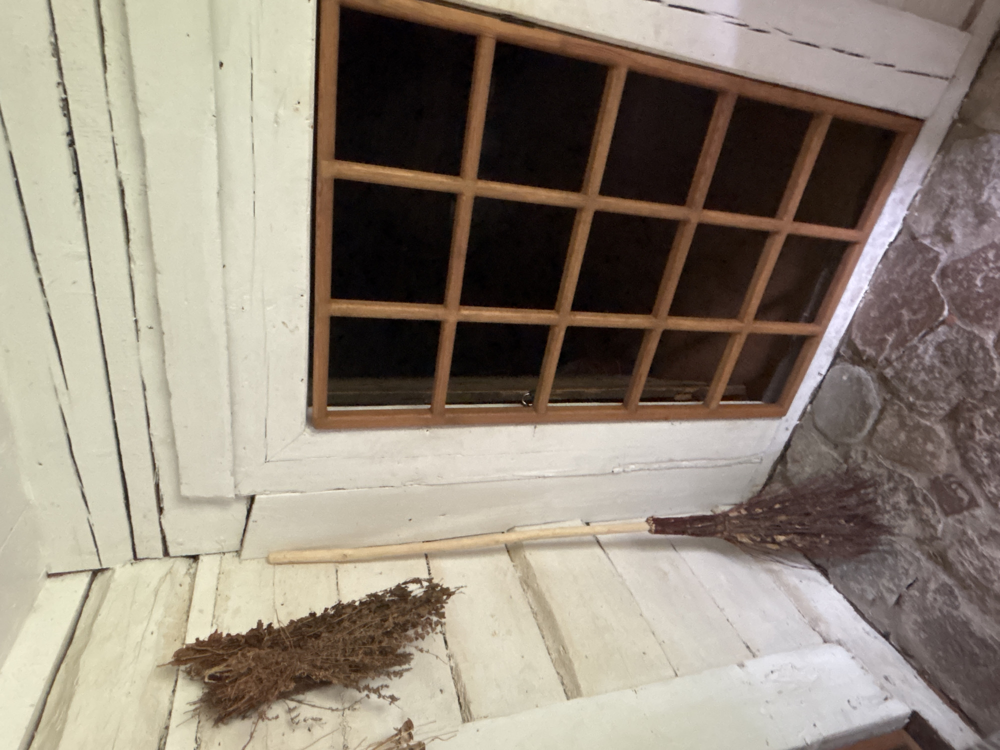 The image depicts a close-up view of a rustic, indoor scene, likely a corner of a room or attic. The primary subjects are a small, handmade broom and a wooden-framed window. The broom, lying on a white wooden floor, has a bundle of dried twigs or straw for bristles attached to a light-colored wooden handle. The floorboards are wide and show signs of age and wear, with visible seams and a slightly uneven surface, all painted white.
To the left of the broom, the wall is constructed of vertical white-painted wooden planks. To the right of the window, a rough, stone wall with irregularly shaped, grey and reddish-brown stones is visible, suggesting a very old or rustic construction.
The window is a key feature, with a dark, unreflective pane divided into a grid of 12 rectangular sections by a light-colored wooden frame. The window is set into a white-painted structure that appears to be part of the wall and ceiling. The overall impression is one of a simple, perhaps historical or miniature, interior space.
There is no discernible activity or event taking place; the scene is static. Notable details include the texture of the broom bristles and the stone wall, the planked construction of the wooden elements, and the grid pattern of the window. The lighting suggests an indoor setting, possibly with natural light coming from the window, though the interior is predominantly dark. There is no visible text in the image. The location context of Sielachowskie, Poland, adds a geographical element to the scene.