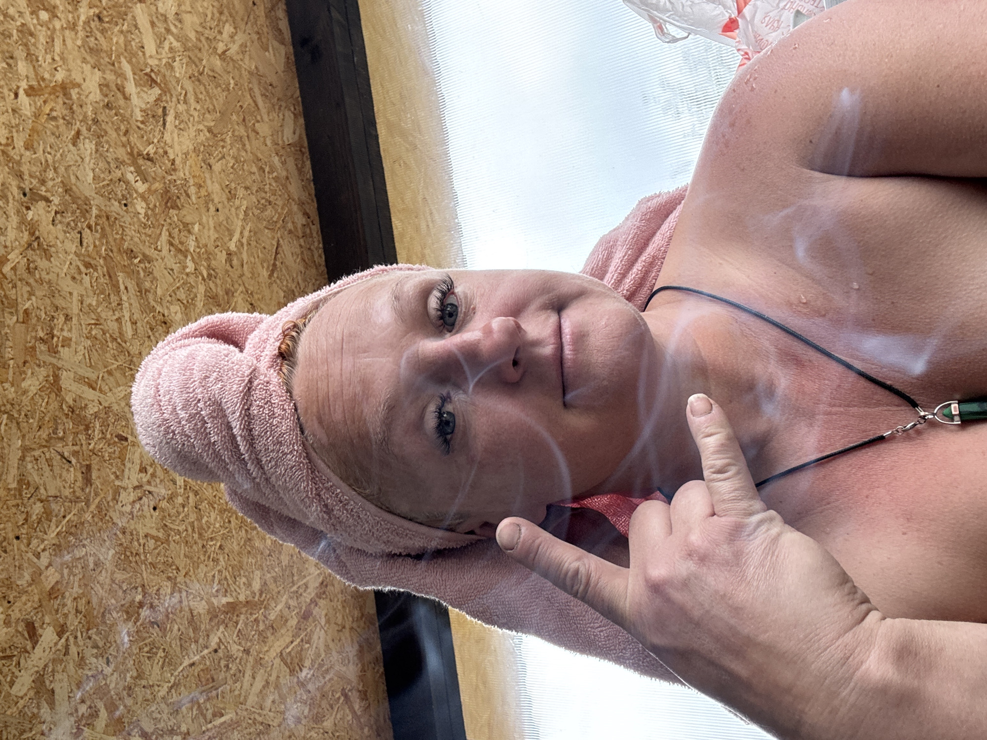 A woman with a towel wrapped around her head is indoors. She is pointing with her index finger. Smoke is visible around her face and upper body. She is wearing a necklace. The background includes what appears to be plywood and a bright white surface. There is no indication in the image that this is in Chaikovsky, Russia.