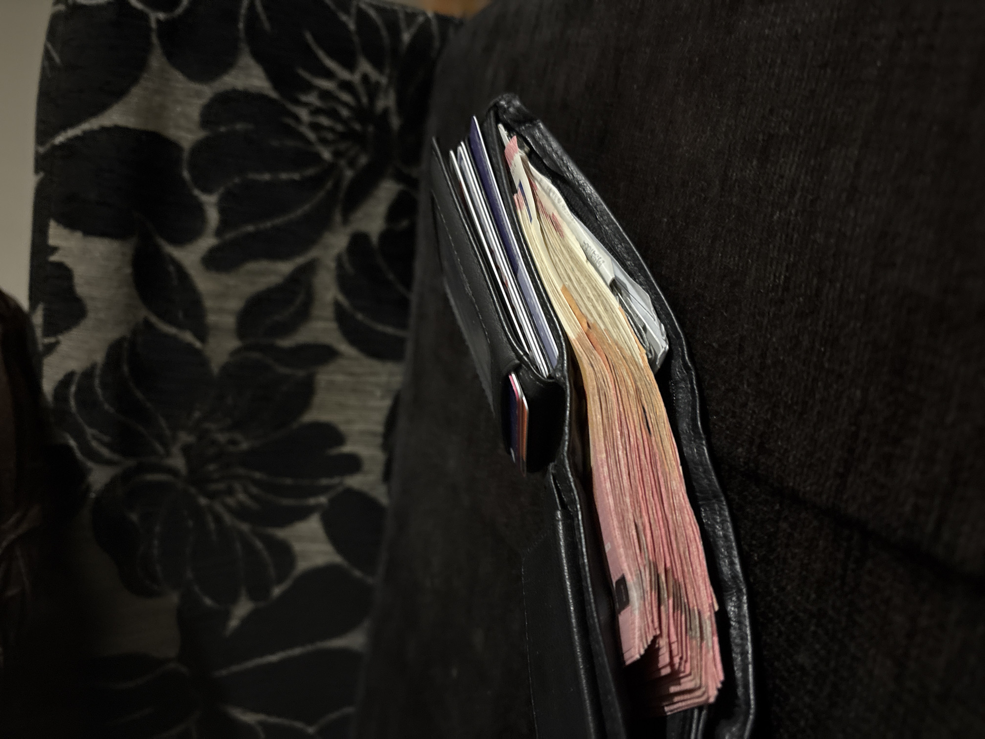 The image shows a black leather wallet stuffed with cash. The currency appears to be euro notes, judging by the pink color. The wallet also contains several cards. The wallet is protruding from the edge of a dark, possibly upholstered surface, suggesting it's placed in a gap or resting against something. To the left is a dark floral patterned fabric, possibly a curtain or part of the upholstery. The location of the image is likely to be Sofia, Bulgaria, since this was the city where the photo was taken.