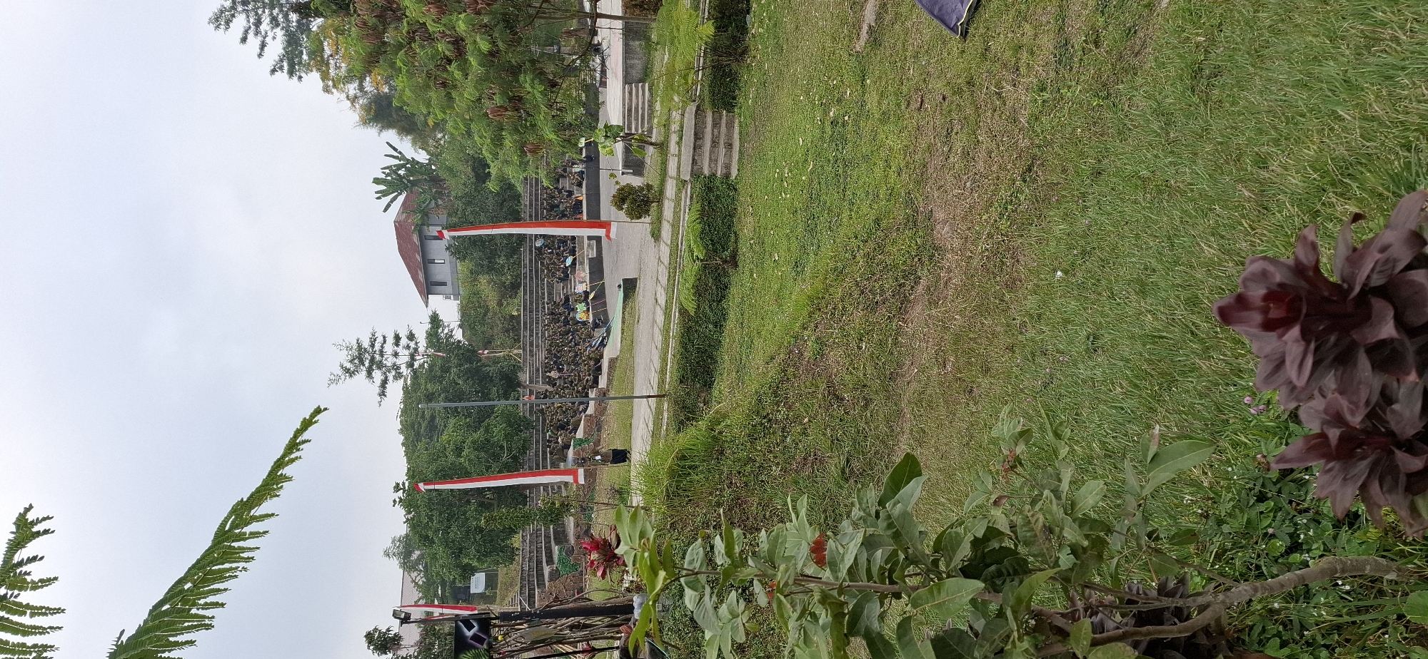 The image depicts an outdoor amphitheater setting, likely in Karang, Indonesia, given the presence of Indonesian flags. In the mid-ground, a large audience, appearing to be predominantly young individuals in uniform khaki-like attire, is seated on tiered stone bleachers. Two tall poles with red and white Indonesian flags are prominently placed near the stage area. The stage itself is a concrete platform. In the foreground, lush green grass covers a sloping area, with a few plants and flowers, including a dark red, leafy plant in the lower right corner. A smaller plant with green leaves is visible in the lower left. Behind the audience and stage, there are buildings, trees, and a stone wall structure. The sky is overcast, suggesting a cloudy day. The overall scene suggests an organized event or gathering, possibly a school or community function.
