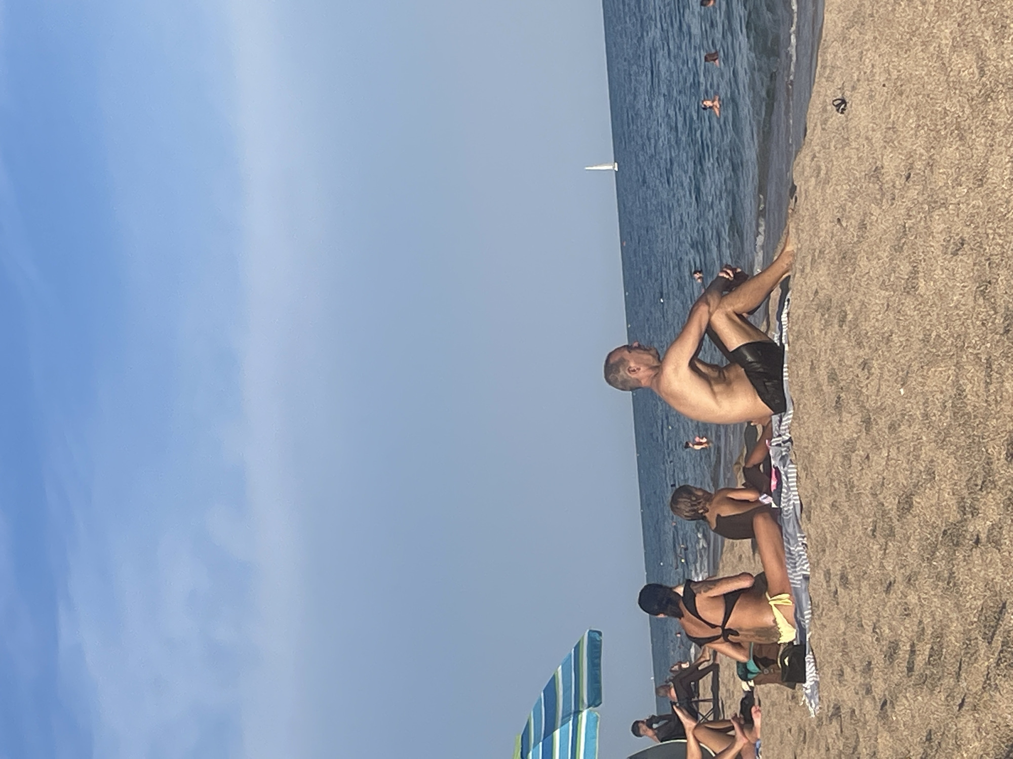 In Canet de Mar, Spain, a sunny beach scene unfolds with various individuals enjoying the day. In the foreground, two people are seated on the sand facing the ocean. A man with a muscular physique, wearing black swim trunks, is sitting with his legs extended. To his left, a woman with a black bikini top and yellow bottoms is also seated, with a young child beside her. Further to the left, partially obscured by an umbrella, another person is visible. The umbrella itself is blue and green striped. The background reveals the calm blue ocean with several people swimming and a white sailboat in the distance. The sky is clear and blue with some wispy clouds.