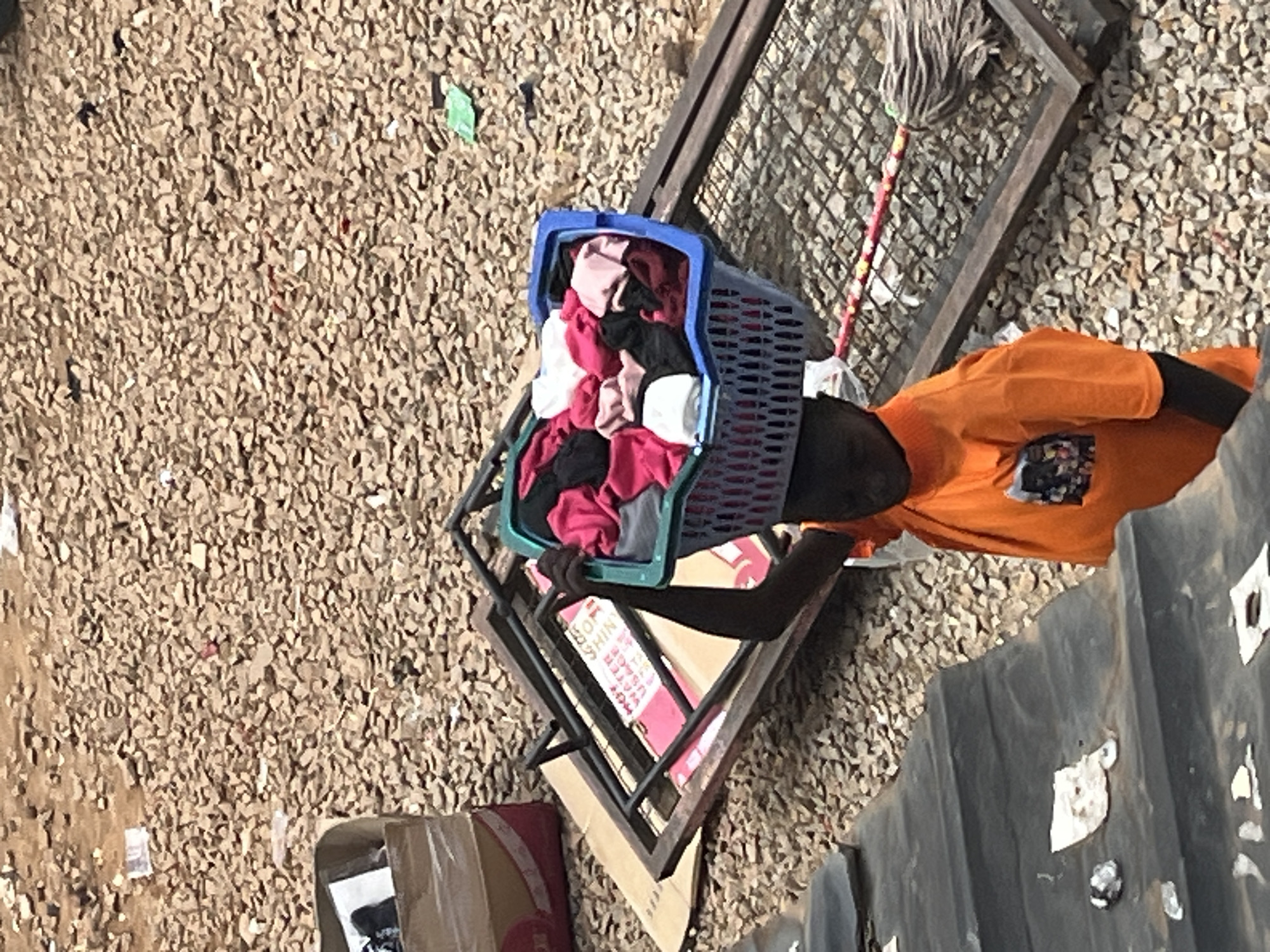 An overhead shot captures a young person, their face obscured, balancing a blue plastic basket overflowing with colorful laundry atop their head. The basket rests on a metal frame, which appears to be part of a larger structure or object. The individual is wearing a bright orange t-shirt. The environment is characterized by a ground covered in light brown gravel, littered with small pieces of debris. To the left, a cardboard box is visible, and further left, a dark, textured surface rises, possibly a rooftop or elevated platform. A mop with a red handle lies on the gravel near the metal frame. The scene is set in a casual outdoor environment, suggesting a street or an open yard in the city of Jos, Nigeria.