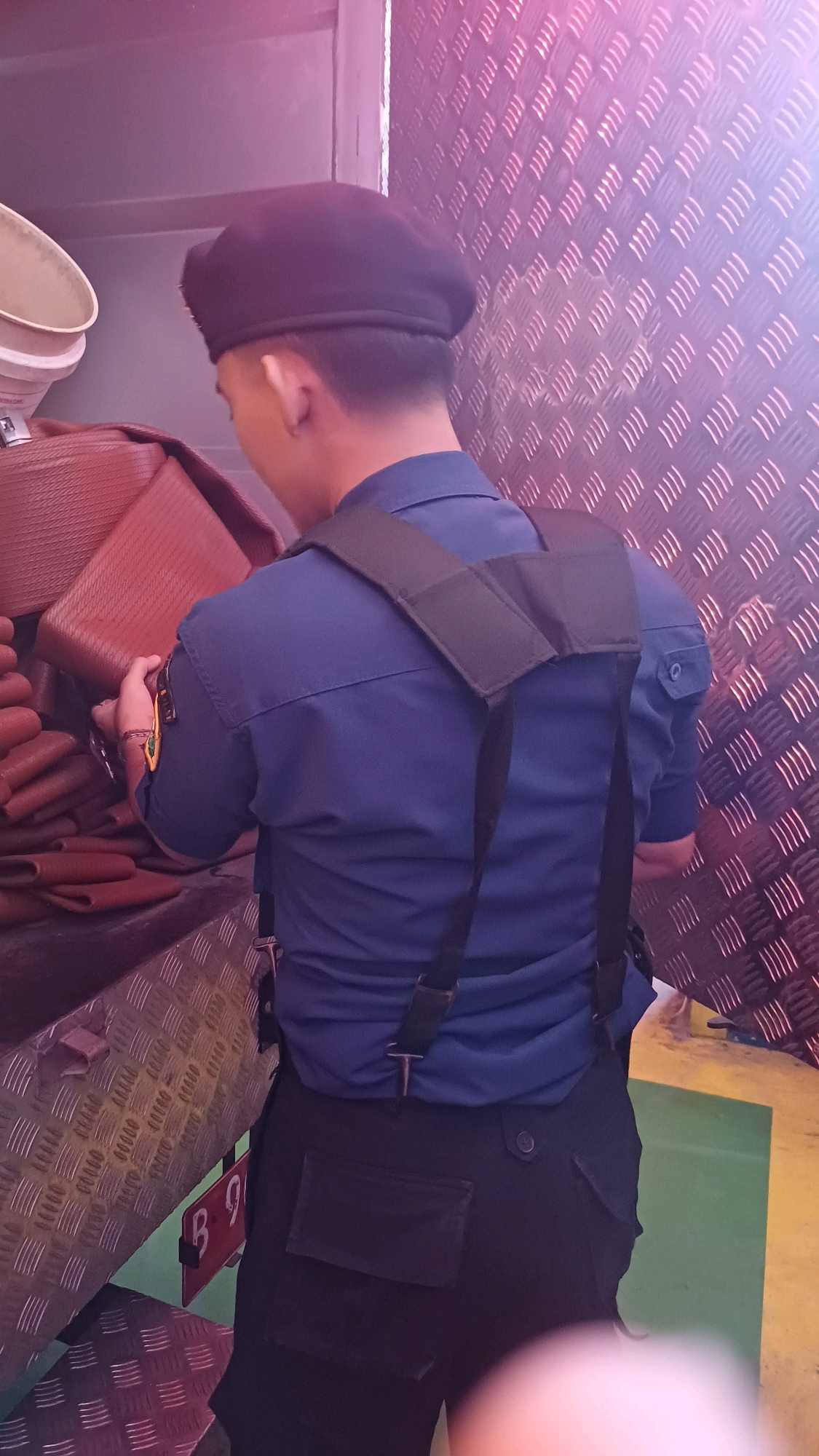 A person wearing a dark beret and a navy blue uniform shirt with black suspenders is seen from behind, handling stacked reddish-brown rectangular objects. The person is standing in a vehicle compartment with a diamond-plate metal wall and floor. A license plate with "B 9" is partially visible on the floor. The scene appears to be in a public transport vehicle or a cargo area within the city of South Jakarta, Indonesia.