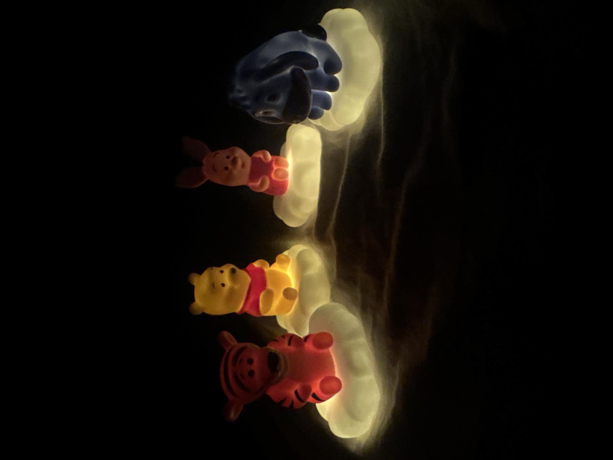 The image shows a vertical arrangement of four Winnie the Pooh character nightlights illuminated in a dark setting. From top to bottom, the characters are Eeyore, Piglet, Winnie the Pooh, and Tigger. Each character is positioned on a cloud-shaped light fixture that emits a soft glow, with the characters themselves also lit up in their respective colors. The background is completely black, which emphasizes the glowing nightlights. There is no indication of location in Votkinsk, Russia, or any other specific place.