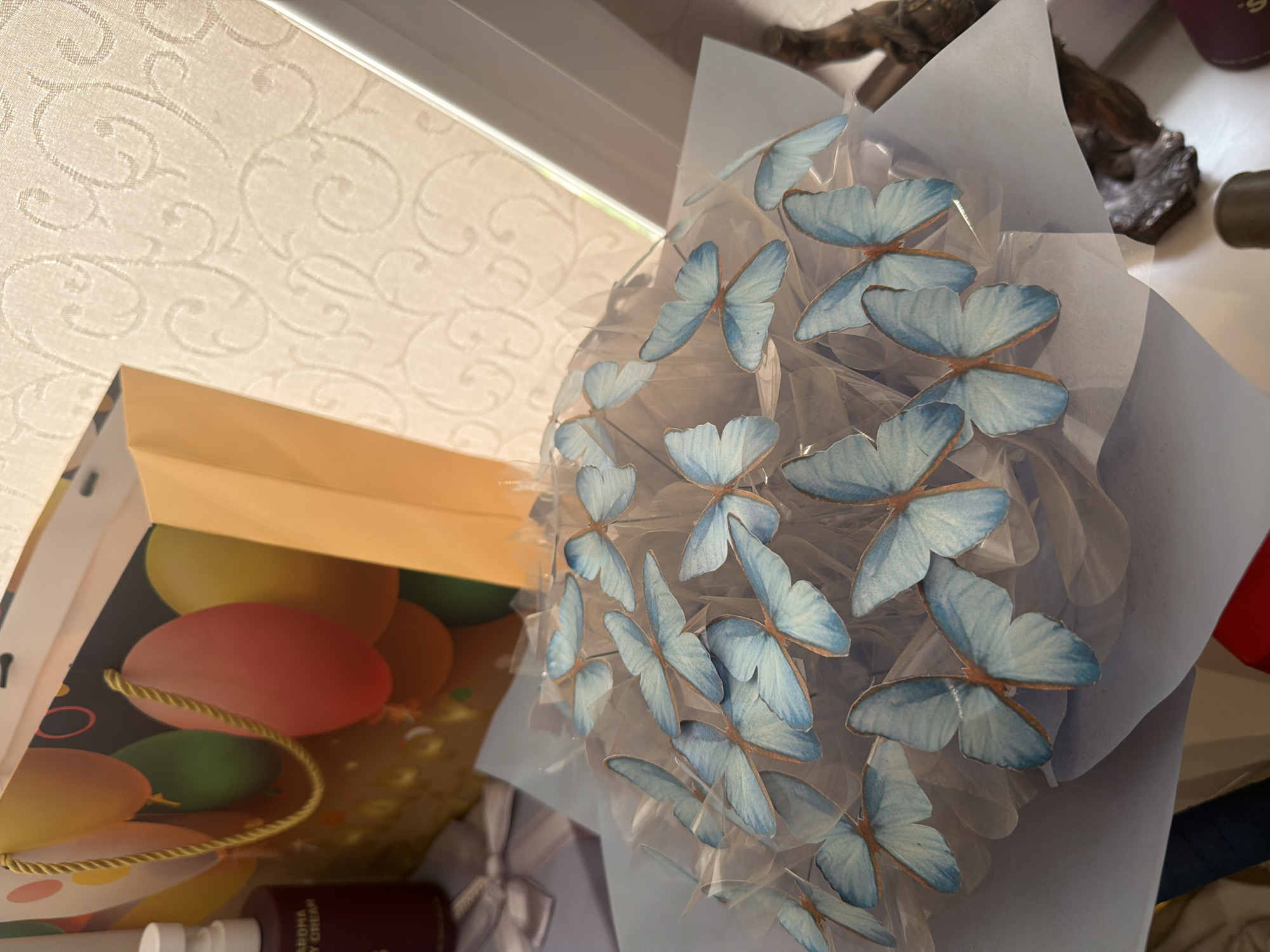 In Dnipro, Ukraine, a bouquet of artificial blue butterflies is arranged in a clear plastic wrapper, placed on a light blue surface. Behind the bouquet, a gift bag with balloons printed on it stands upright. To the left of the gift bag, a dark maroon bottle with a white cap, labeled "AROMA BODY CREAM," is visible. A pale pink ribbon is tied in a bow to the left of the bottle. The background features a textured, cream-colored surface with a swirling pattern, suggesting it may be wallpaper or fabric. A window with blinds is visible on the right side of the image, letting in natural light.